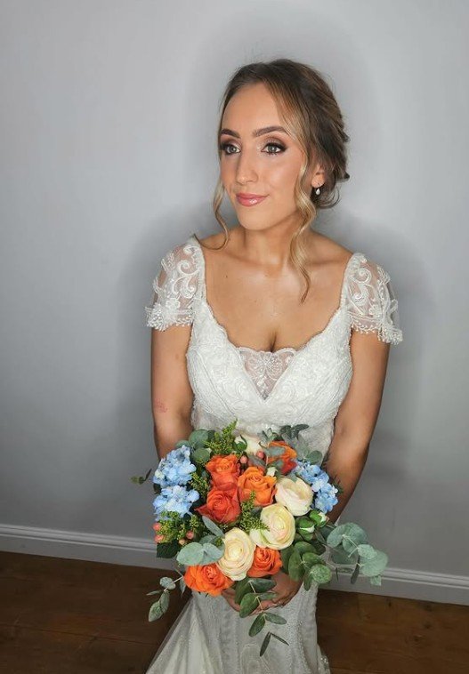 A bride in a white lace wedding dress holding a bouquet of flowers with orange roses, white roses, blue flowers, and green leaves, standing against a plain gray wall.