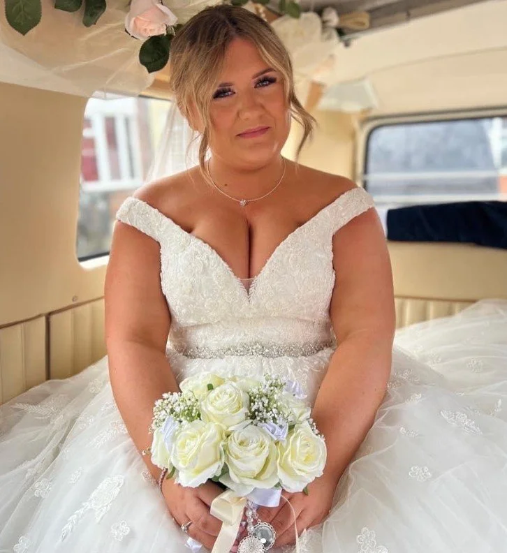 A woman in a white wedding dress holding a bouquet of white roses, sitting inside a vehicle with a window showing buildings outside.
