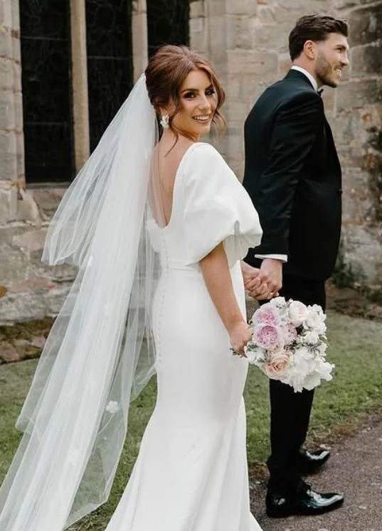 Bride and groom holding hands outdoors, bride wearing a white wedding dress with a long veil and holding a bouquet of pink and white flowers, groom in a black tuxedo, in front of a stone building.
