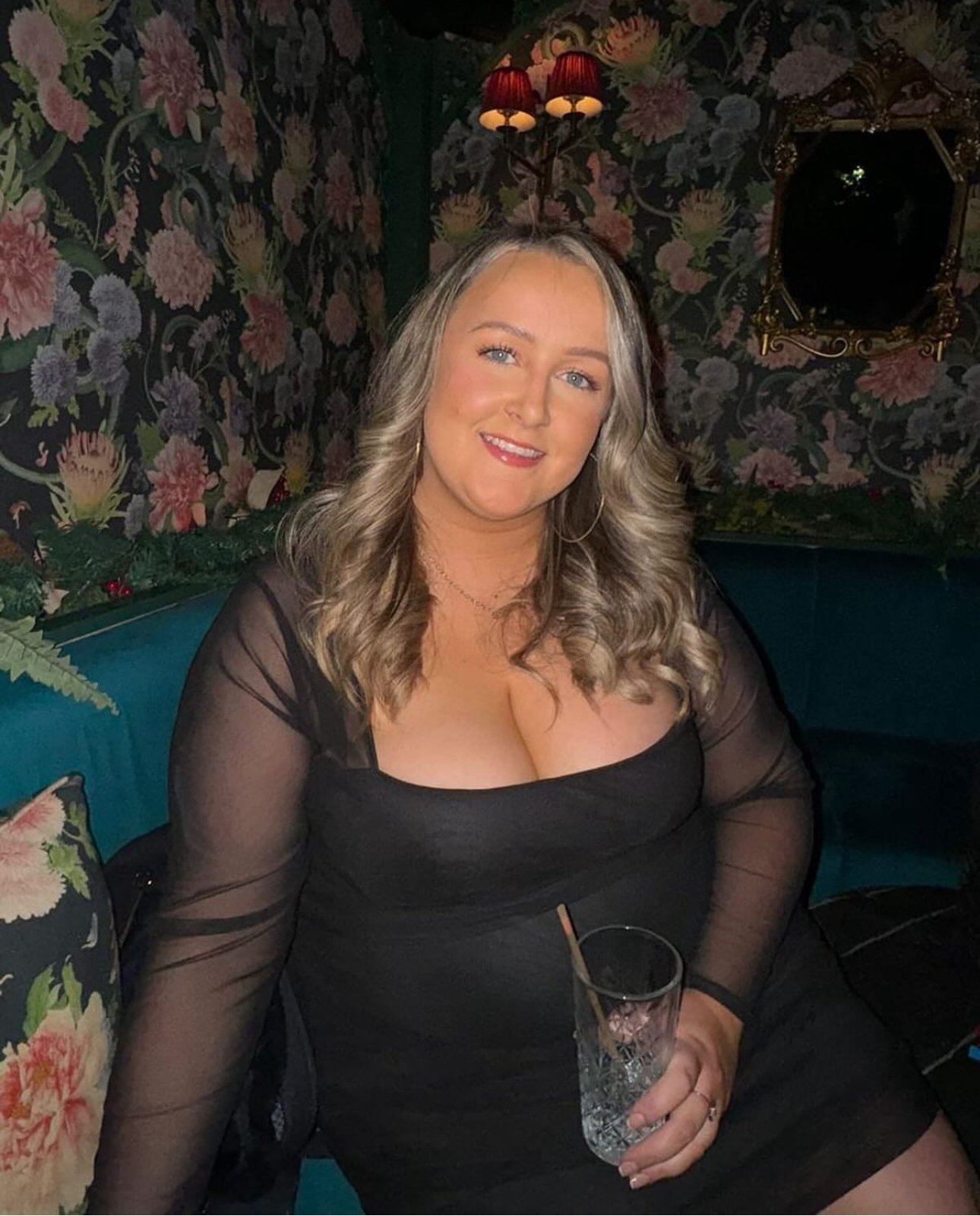 A woman with blonde, wavy hair, wearing a black dress with sheer sleeves, smiling while holding a glass with a straw. She is sitting on a teal sofa in a room with floral wallpaper and decorative lighting.