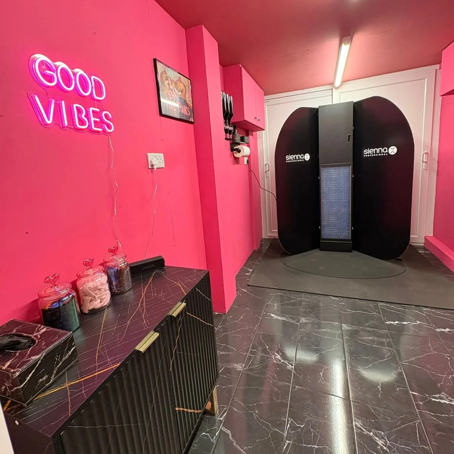 A pink wall with a neon sign that reads 'Good Vibes', a framed picture, a black marble cabinet with jars.