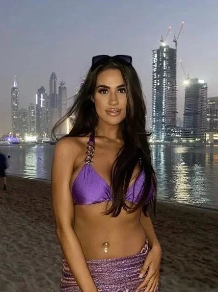 A woman with long dark hair wearing a purple bikini top with embellished straps, standing on a beach at dusk with a city skyline with tall buildings and construction cranes in the background.