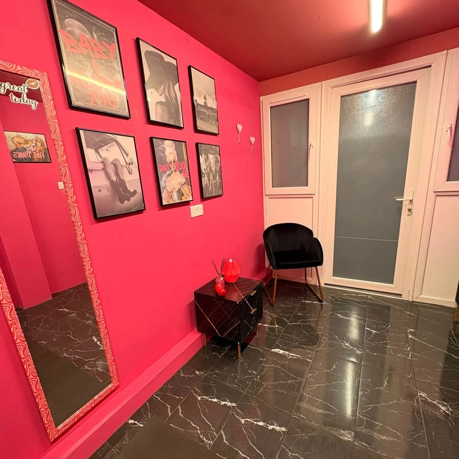 A pink living room corner with black marble floor, a black chair, a small black side table with a red lamp, a wall decorated with framed posters, a large mirror on the left, a door with frosted glass windows, and small wall decorations.