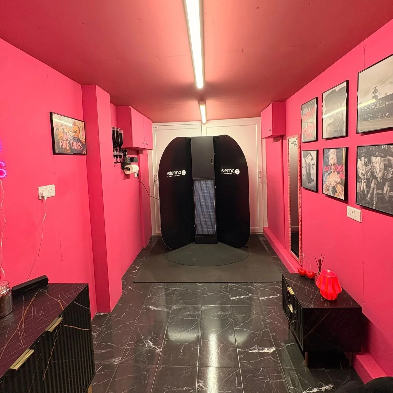 Solihull Spray Tans by Ivy treatment room in Solihull - Pink-coloured room with black marble flooring, featuring a black spray tanning booth and framed posters on the pink walls.