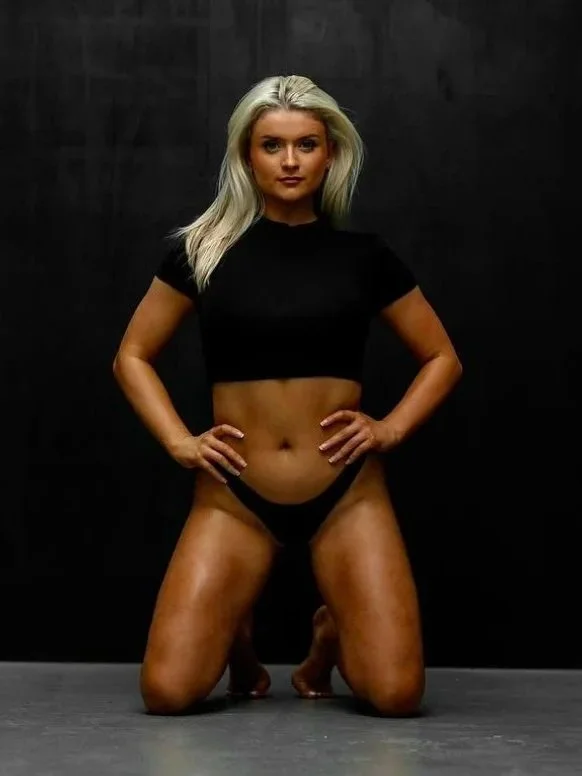 A woman with platinum blonde hair kneeling on the floor with her hands on her hips, wearing a black crop top and black underwear, against a dark background.