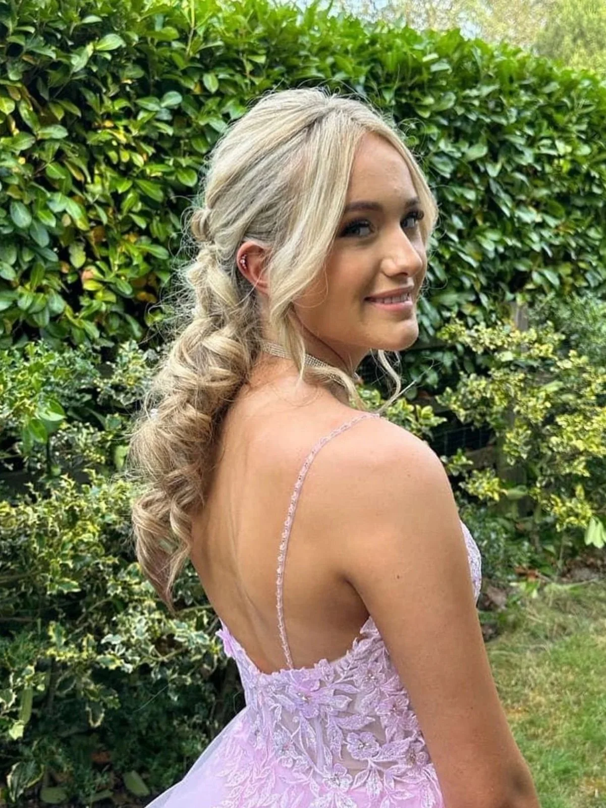A woman with blonde hair styled in loose curls, wearing a light pink embroidered dress with thin straps, standing outdoors in front of green bushes and foliage, smiling.
