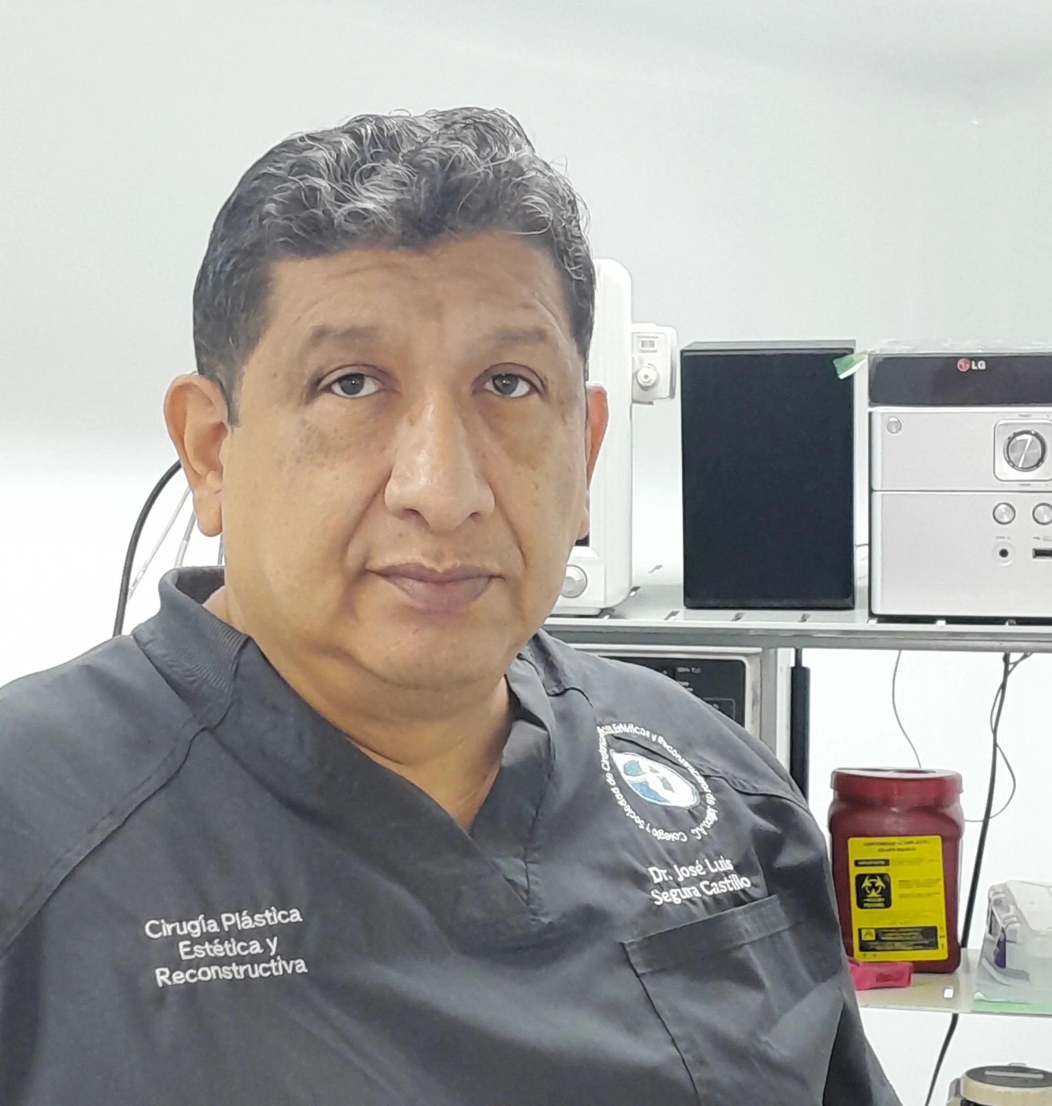 Hombre con short gris oscuro y pálido, con cabello rizado y canoso, cabina de computadoras en un ambiente clínico o de oficina. En la parte superior, en la parte izquierda, se ven monitores y en el centro, un electrocardiograma. La imagen está centrada en la cabeza y torso del hombre.
