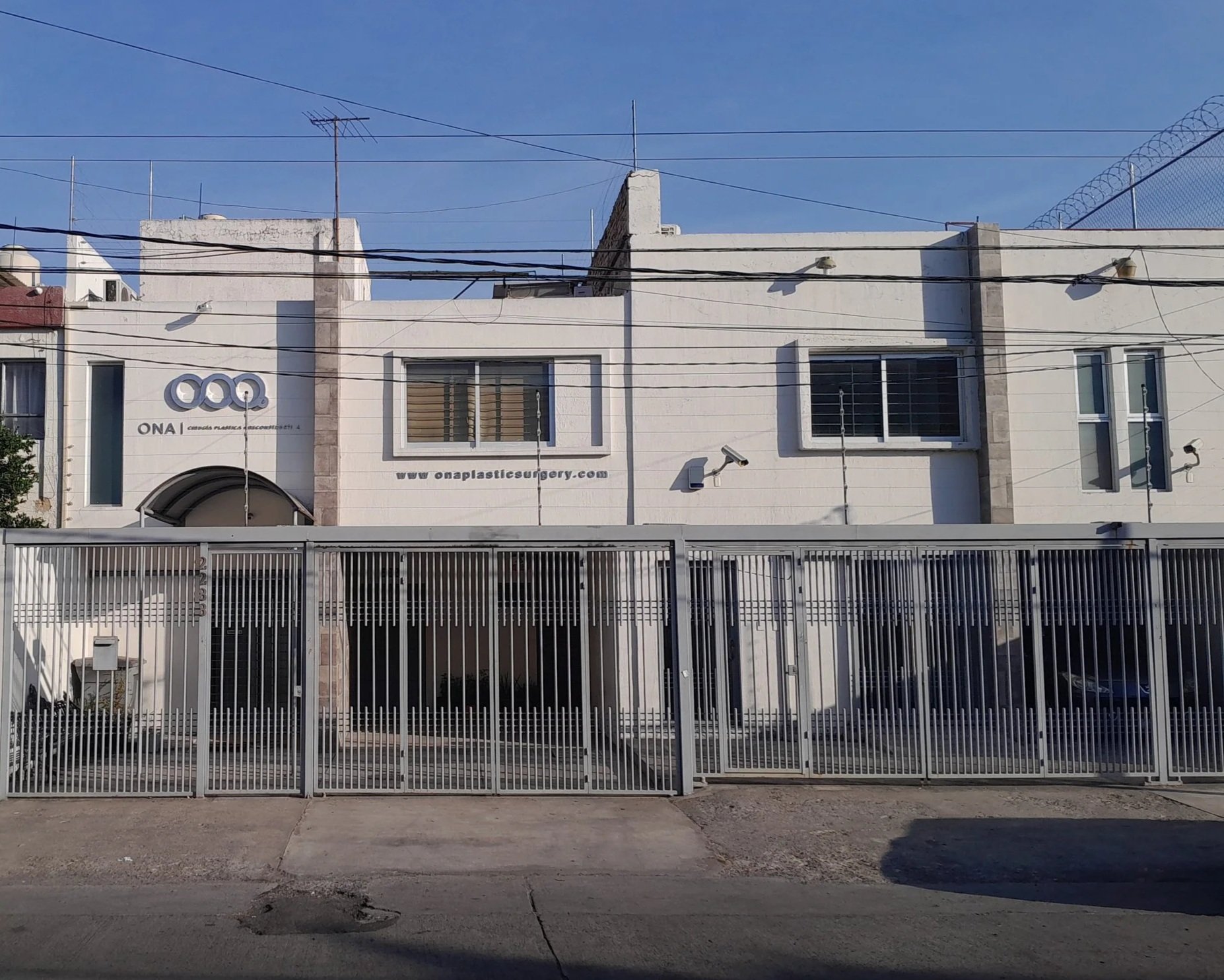 Vista frontal de un edificio de color blanco, con ventanas y un cartel que dice 'ONA cirugía plástica y reconstructiva', con un portón de rejas metálicas en el frente, y cables eléctricos en el cielo azul claro.