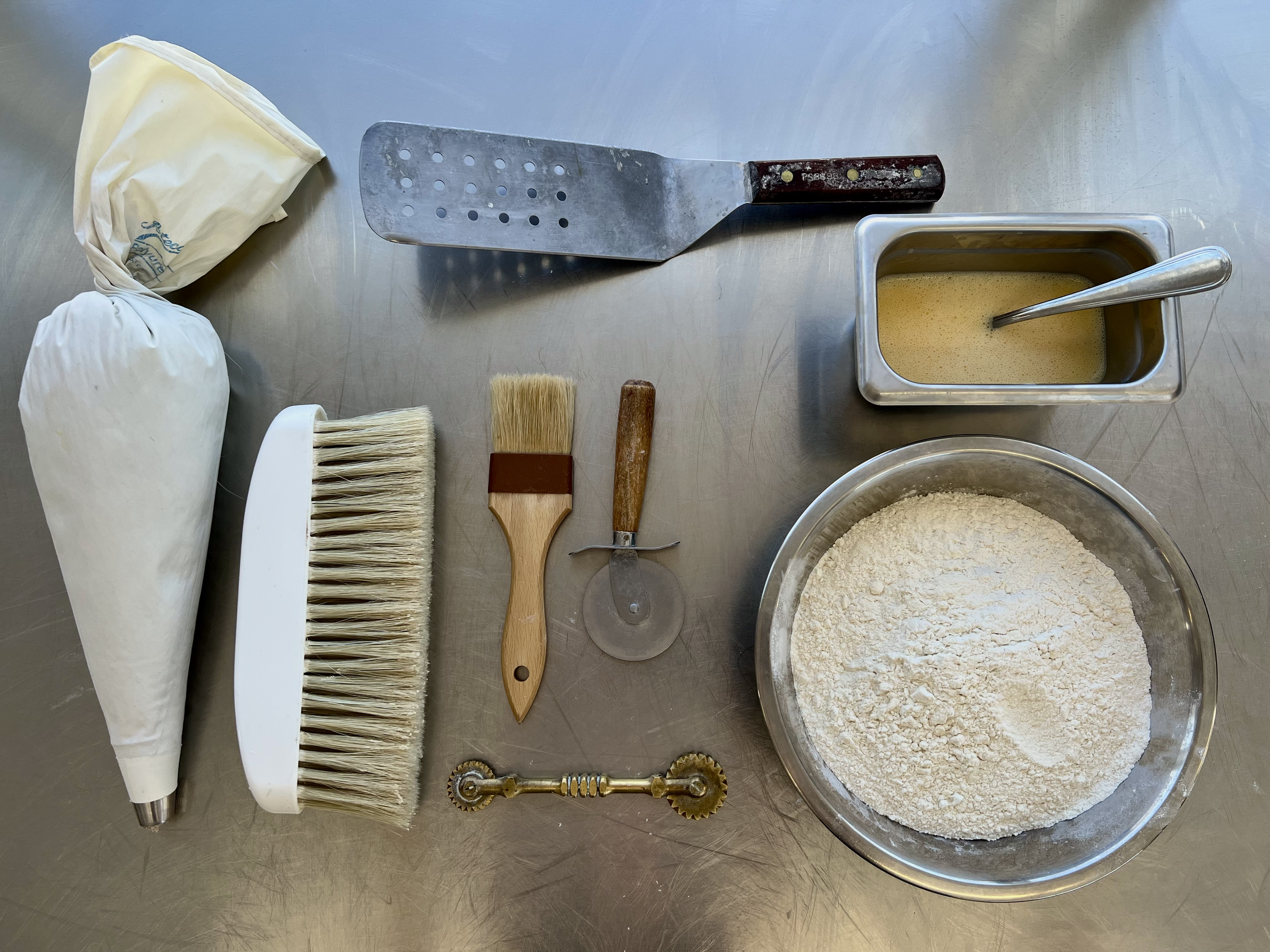 Assorted baking tools and ingredients on a stainless steel surface, including a piping bag, dough scraper, pastry brush, pizza cutter, two pastry brushes, a pasta roller, a metal container with yellow sauce, a large mixing bowl of flour, and a contai