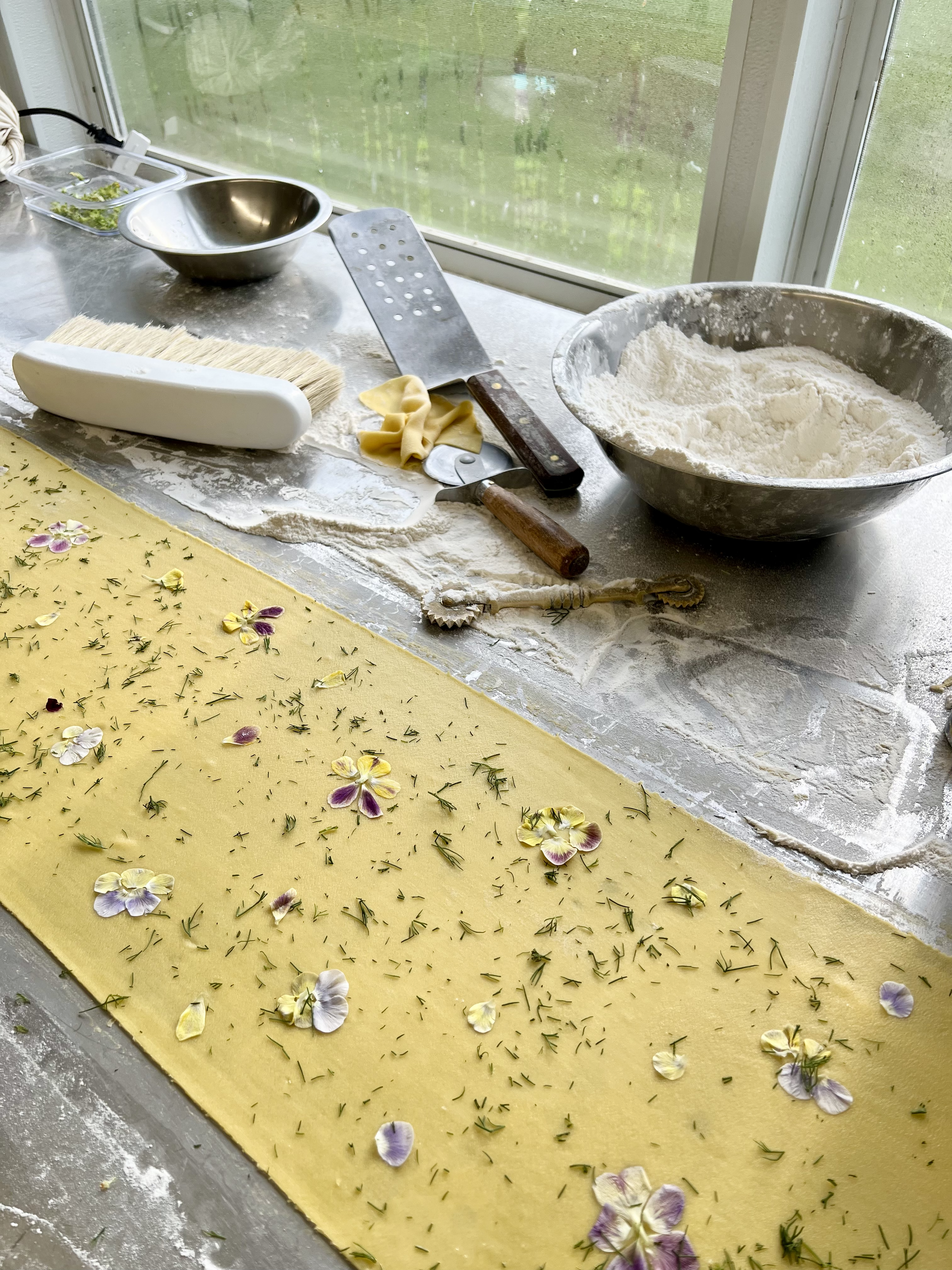 A baking workspace with a yellow dough sheet decorated with edible flowers and herbs, and various baking tools on a floured countertop near a window.