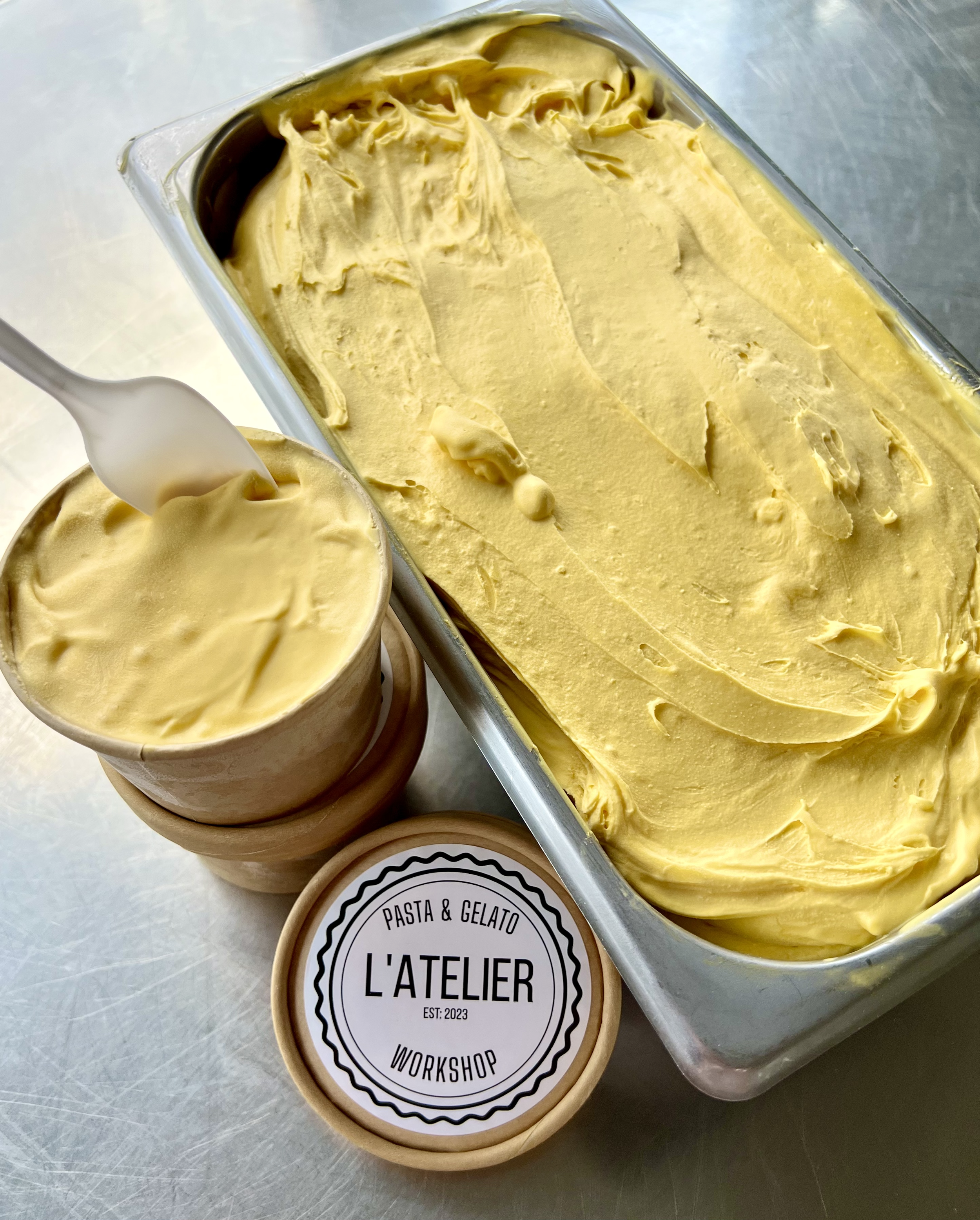 A large container of yellow gelato or ice cream with a smooth surface, a cup of the same yellow gelato, and a labeled container from L'ATELIER with the words 'PASTA & GELATO WORKSHOP'.