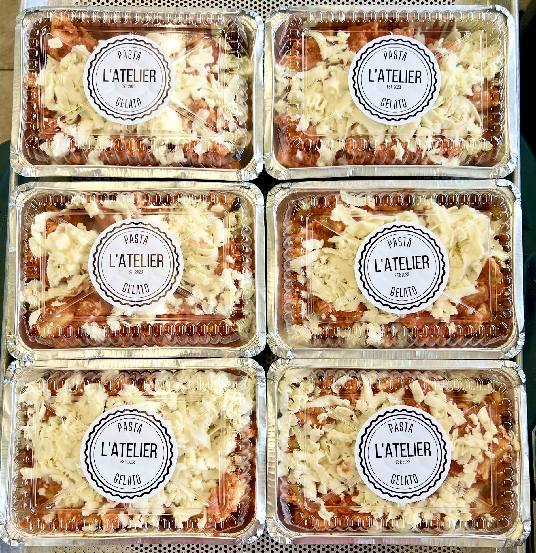 Six containers of pasta with shredded cheese on top, labeled L'ATELIER GELATO, arranged in two rows of three.