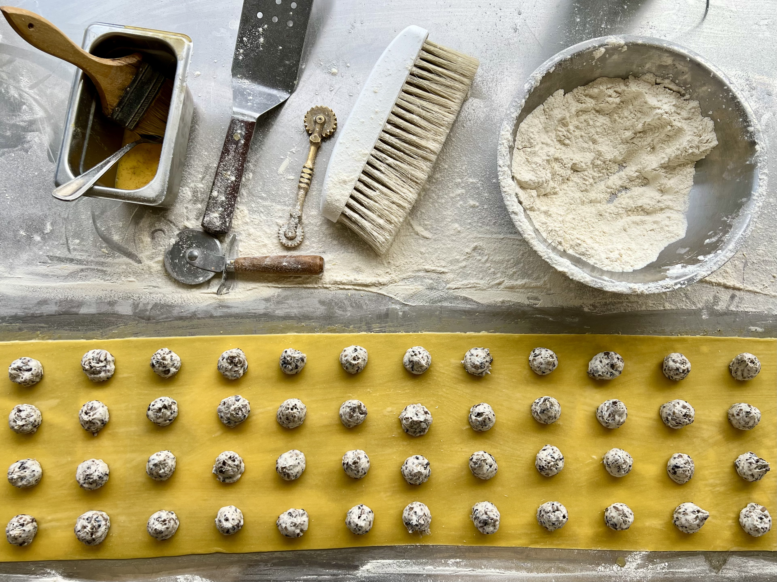 Baking scene with cookie dough portions on yellow parchment paper, flour, rolling pin, bench scraper, flour sifter, brush, and pastry cutter on a floured surface.