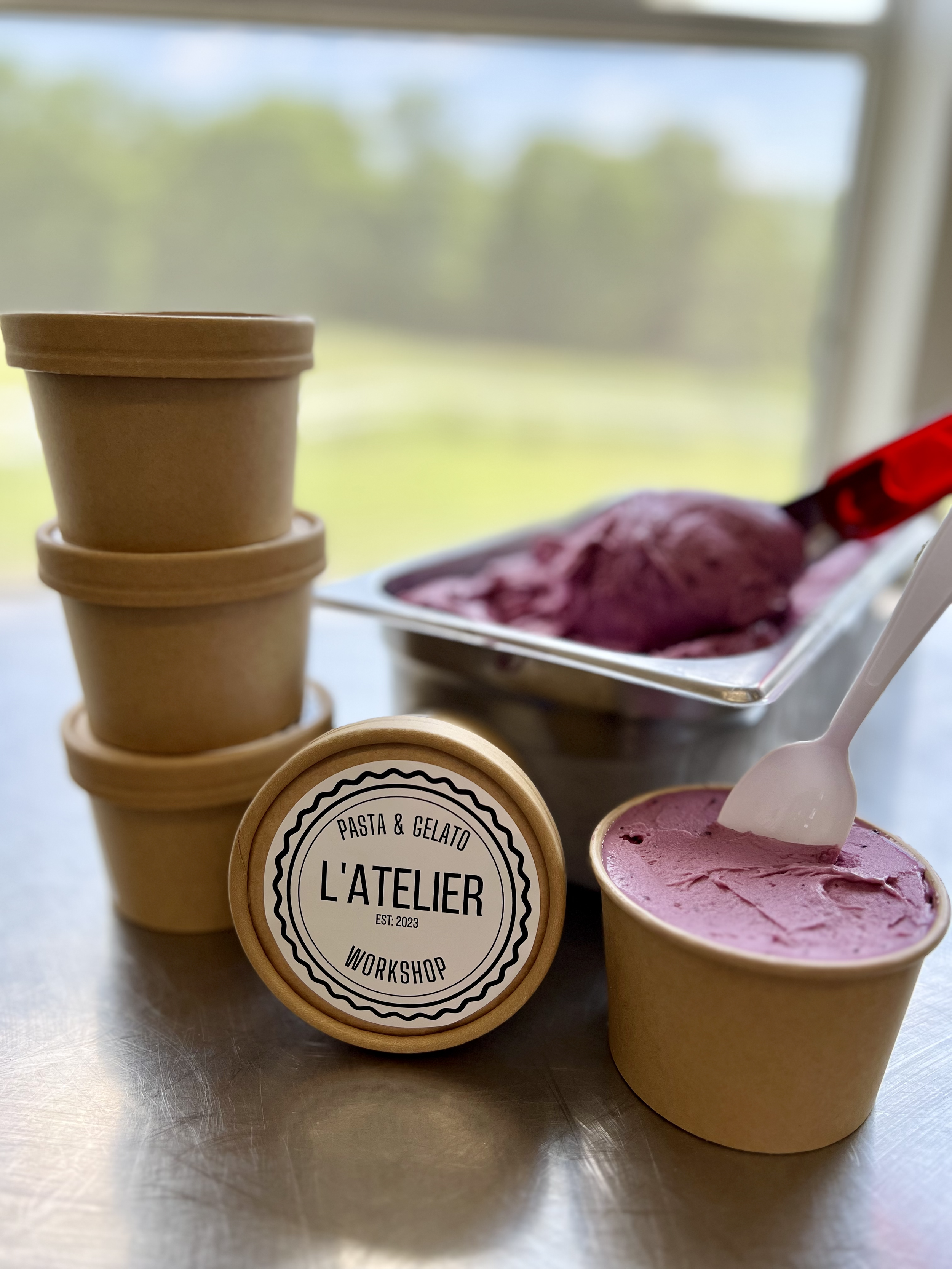 Three stacked brown paper cups, a container with purple gelato, and a cup of pink gelato with a white spoon on a metal surface, with a window in the background.