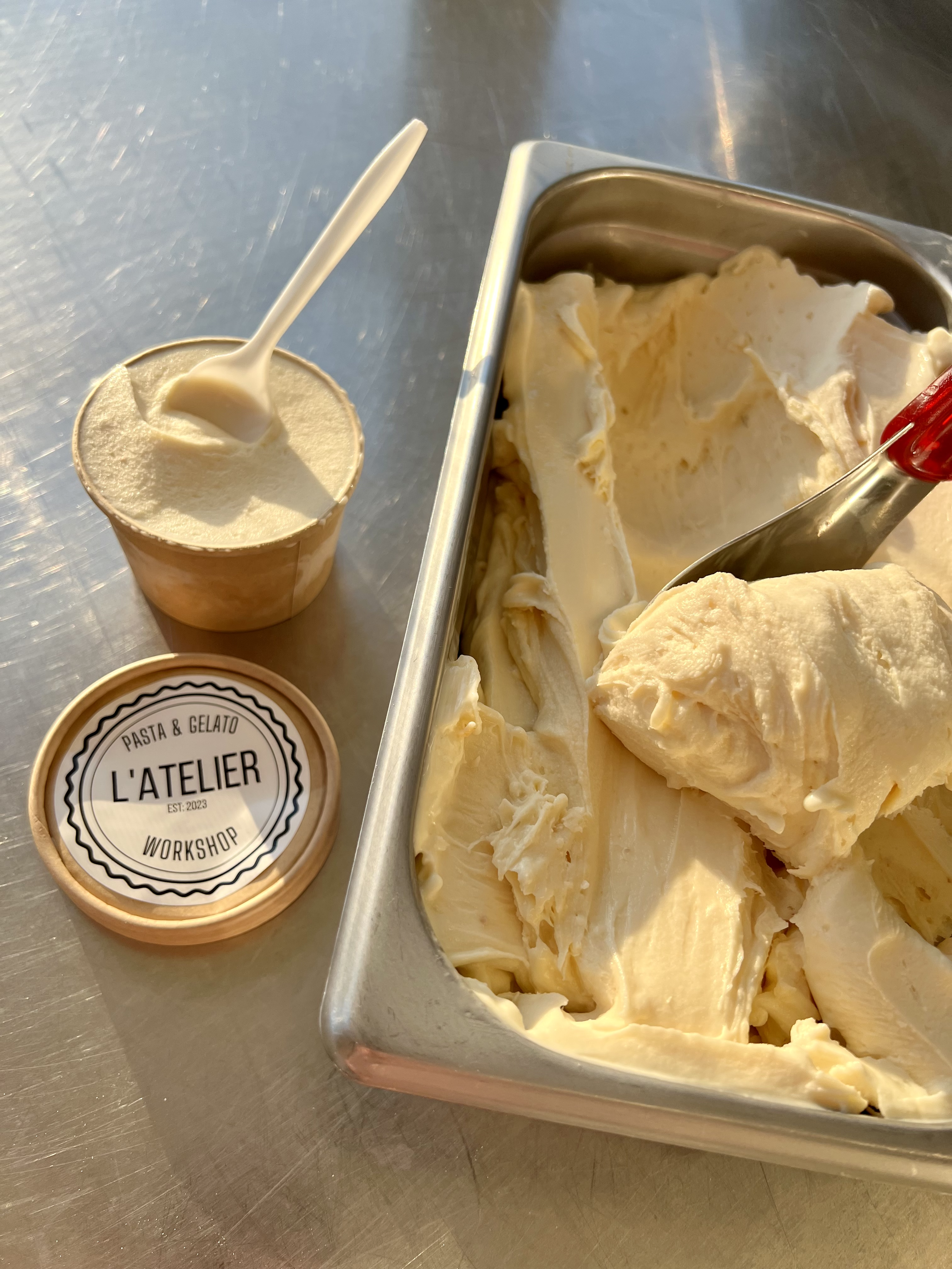Various flavors of gelato on a stainless steel table, with a cup of soft serve ice cream and a container of creamy gelato that has a scoop partially removed.