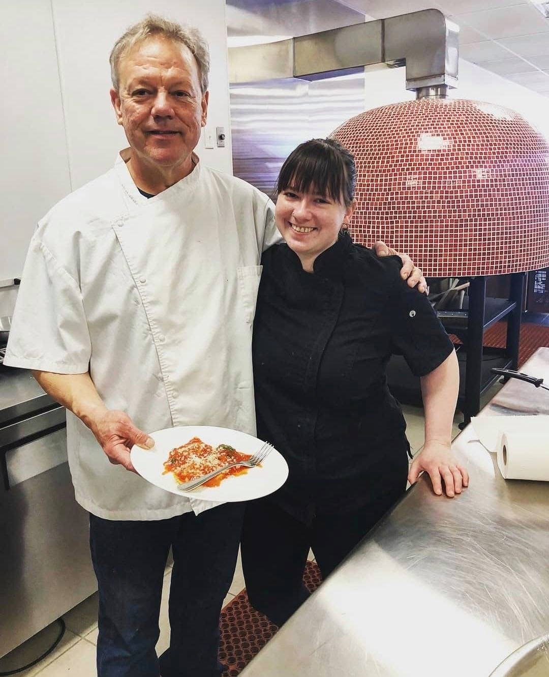 A male chef and a female chef smiling and posing in a commercial kitchen. The male chef is holding a plate of food with sauce and grated cheese. A large pizza oven is visible in the background.