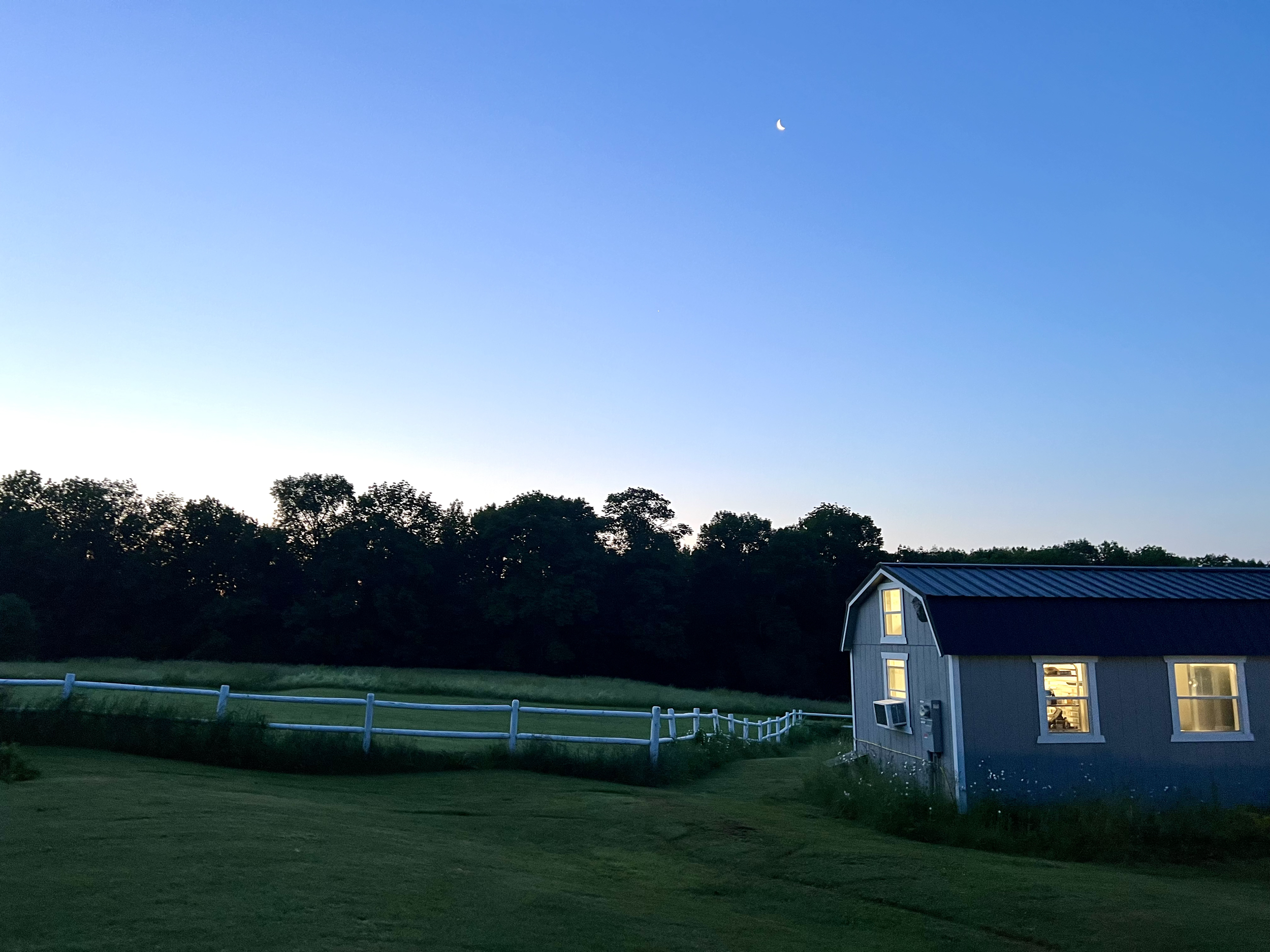 A rural scene at dusk with a small blue house with lights on, a white fence running across a grassy field, and a clear sky with a visible crescent moon.