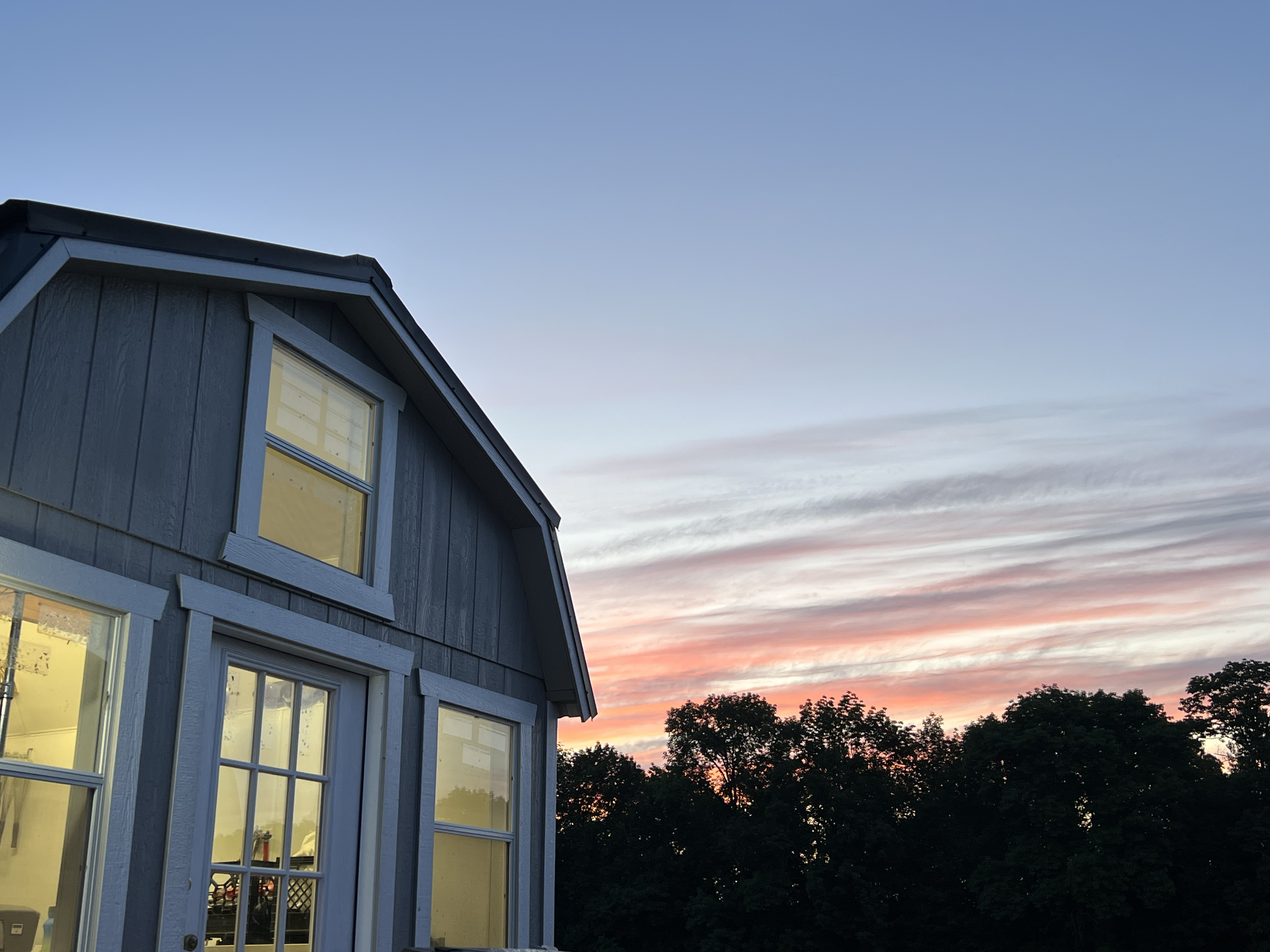 A blue house with a curved roof and large windows at sunset, with dark trees against a sky with pink and orange clouds.