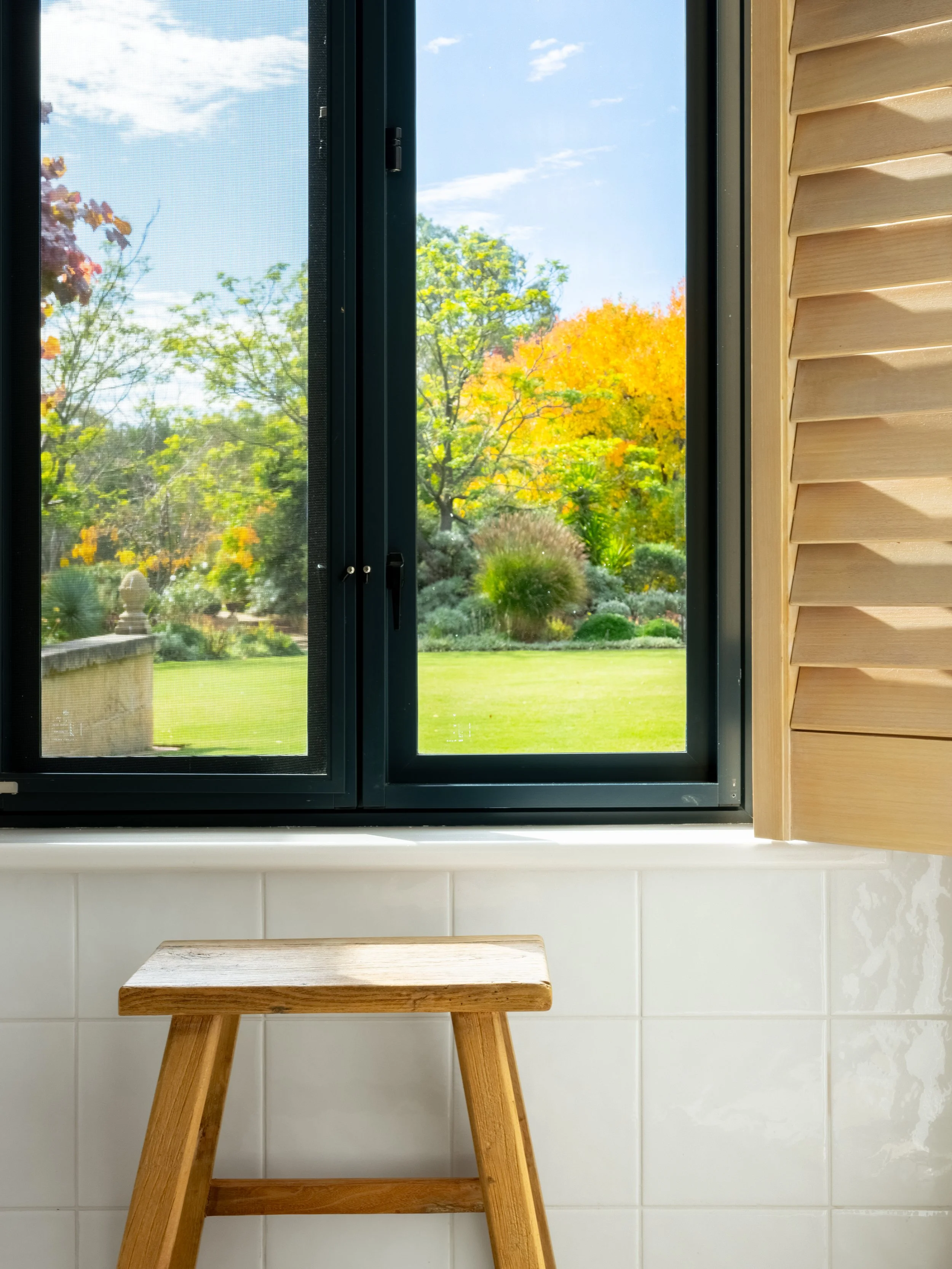 A kitchen windowsill with a small wooden stool in front, looking out to a colorful autumn garden with trees with yellow, orange, and green leaves, under a blue sky with some clouds.