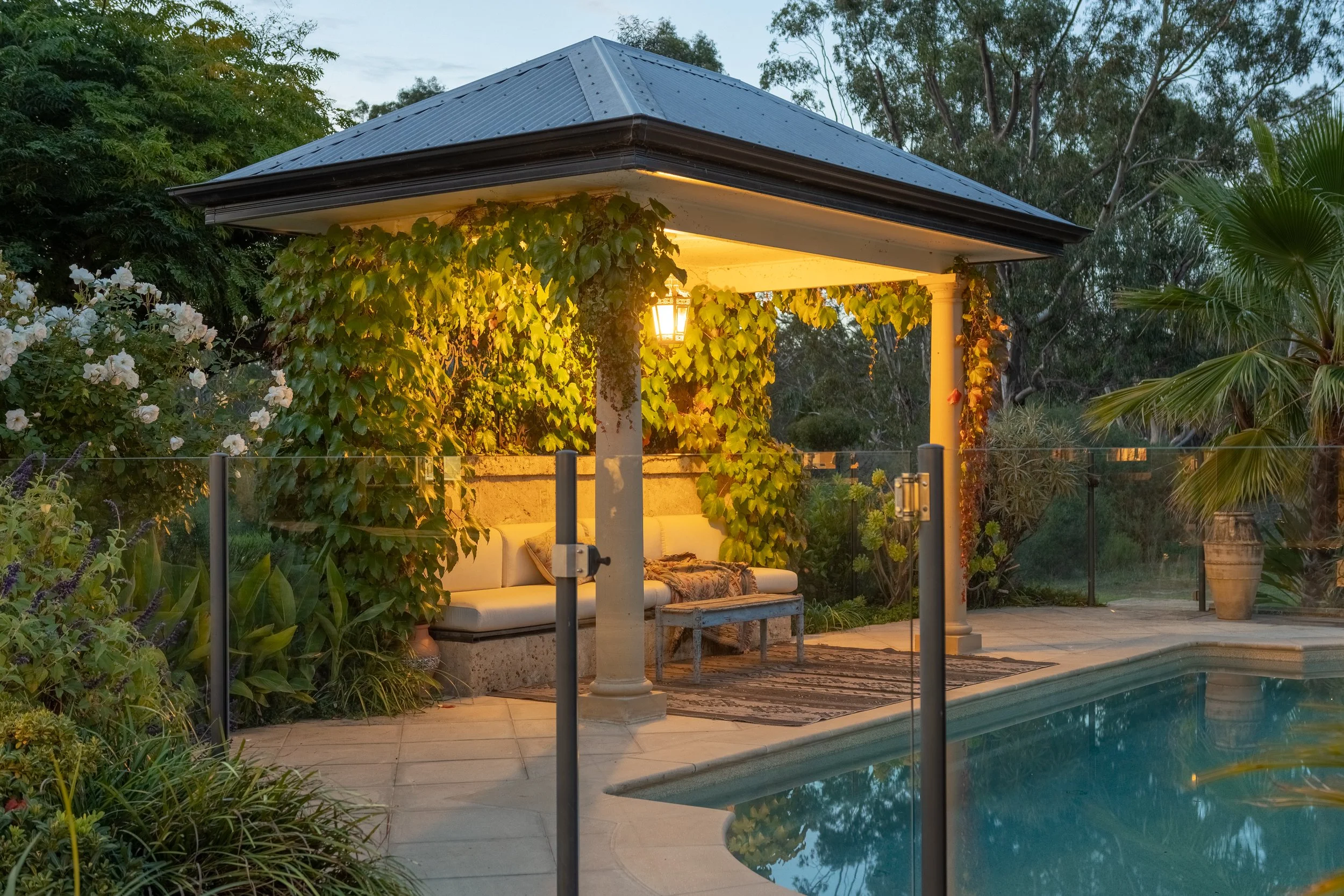 A backyard with a covered patio, hanging ivy, and outdoor seating near a swimming pool. There is a lantern light hanging from the ceiling of the patio, which has a yellow glow, and trees around the area.