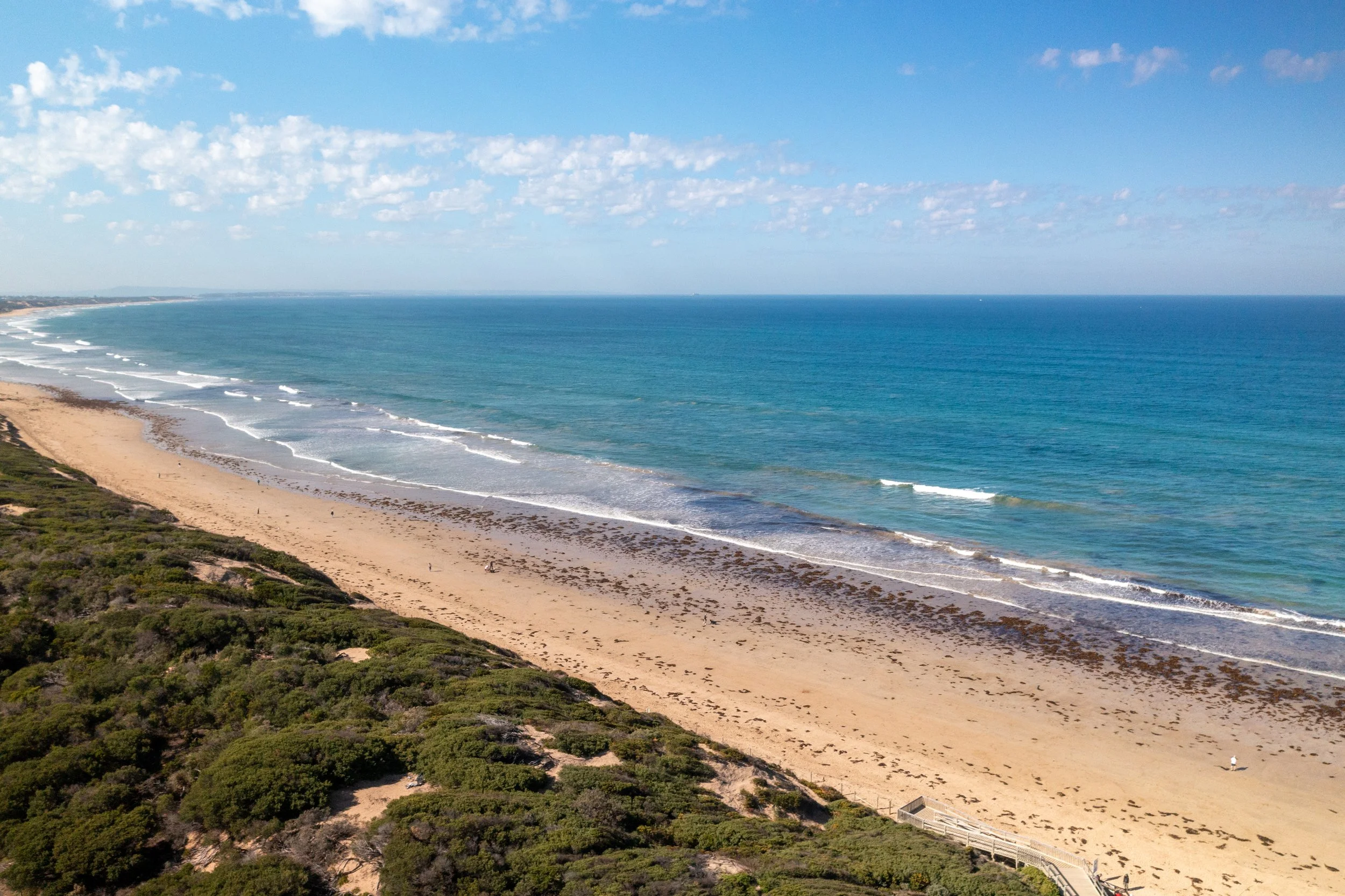 Aerial view of a sandy beach with ocean waves and a boardwalk, bordered by green shrubbery and clear blue sky with scattered clouds.
