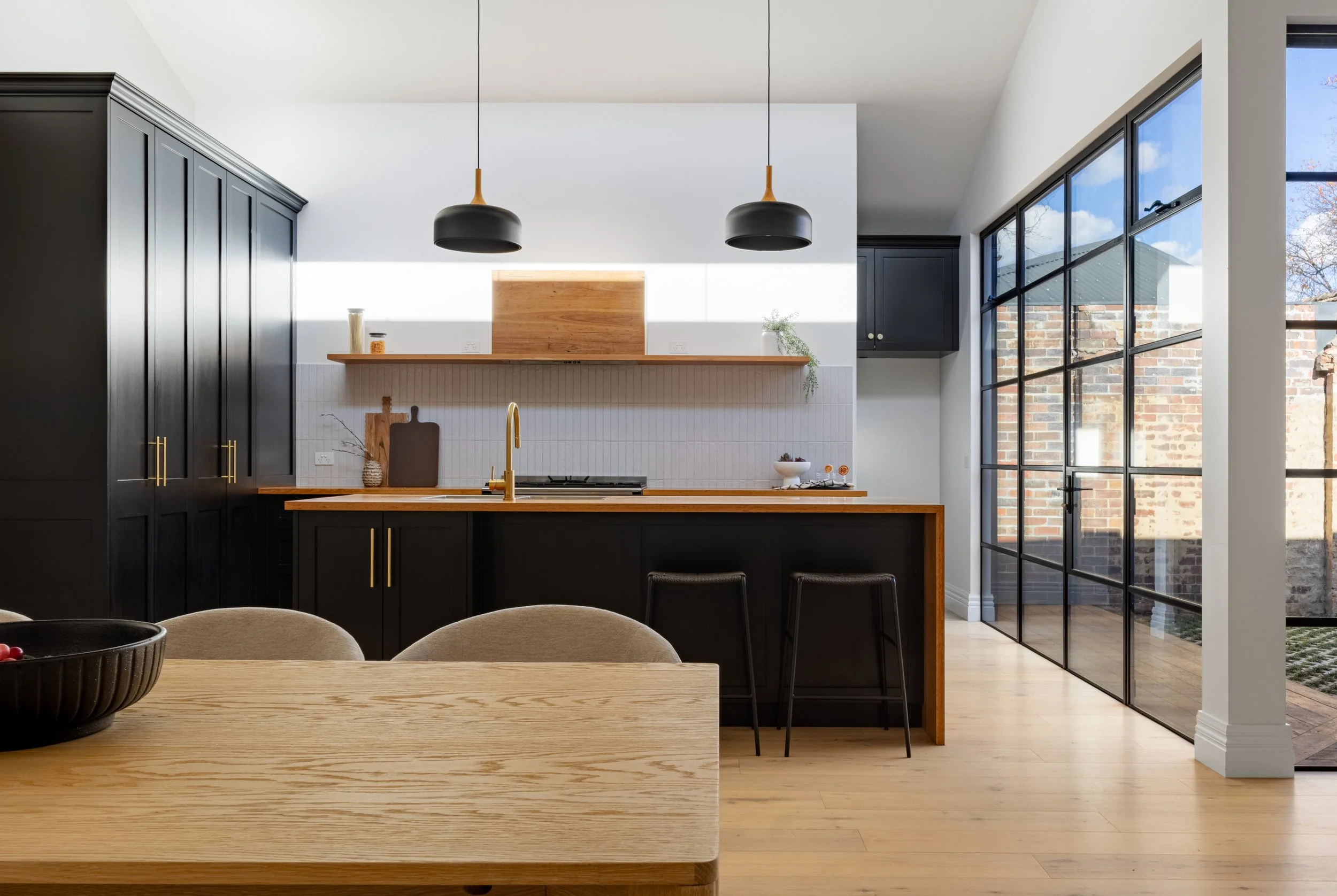 Modern kitchen with black cabinets, wooden countertops, pendant lights, and large glass window showing outdoor brick wall and sky.