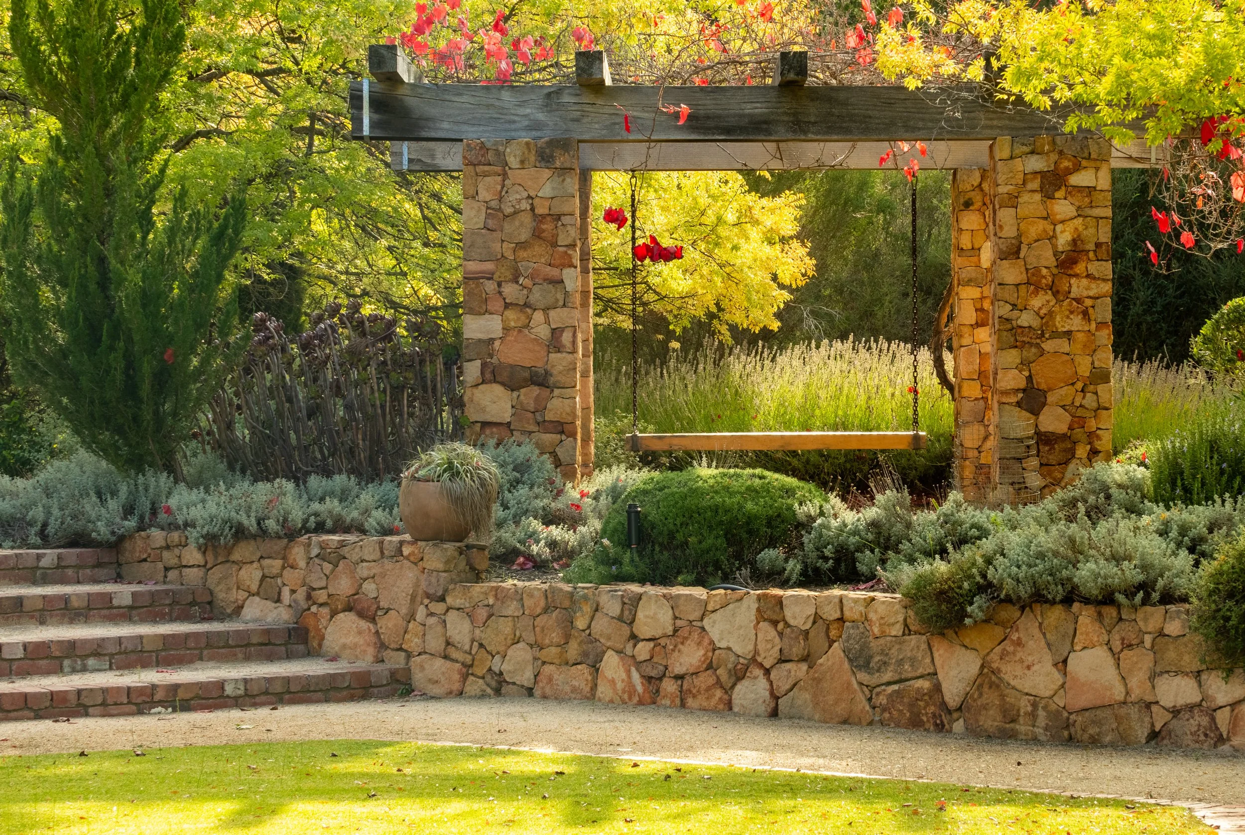 A stone and wood structure with a wooden swing hanging from the top beam, set in a lush garden with trees, shrubs, and flowering plants.