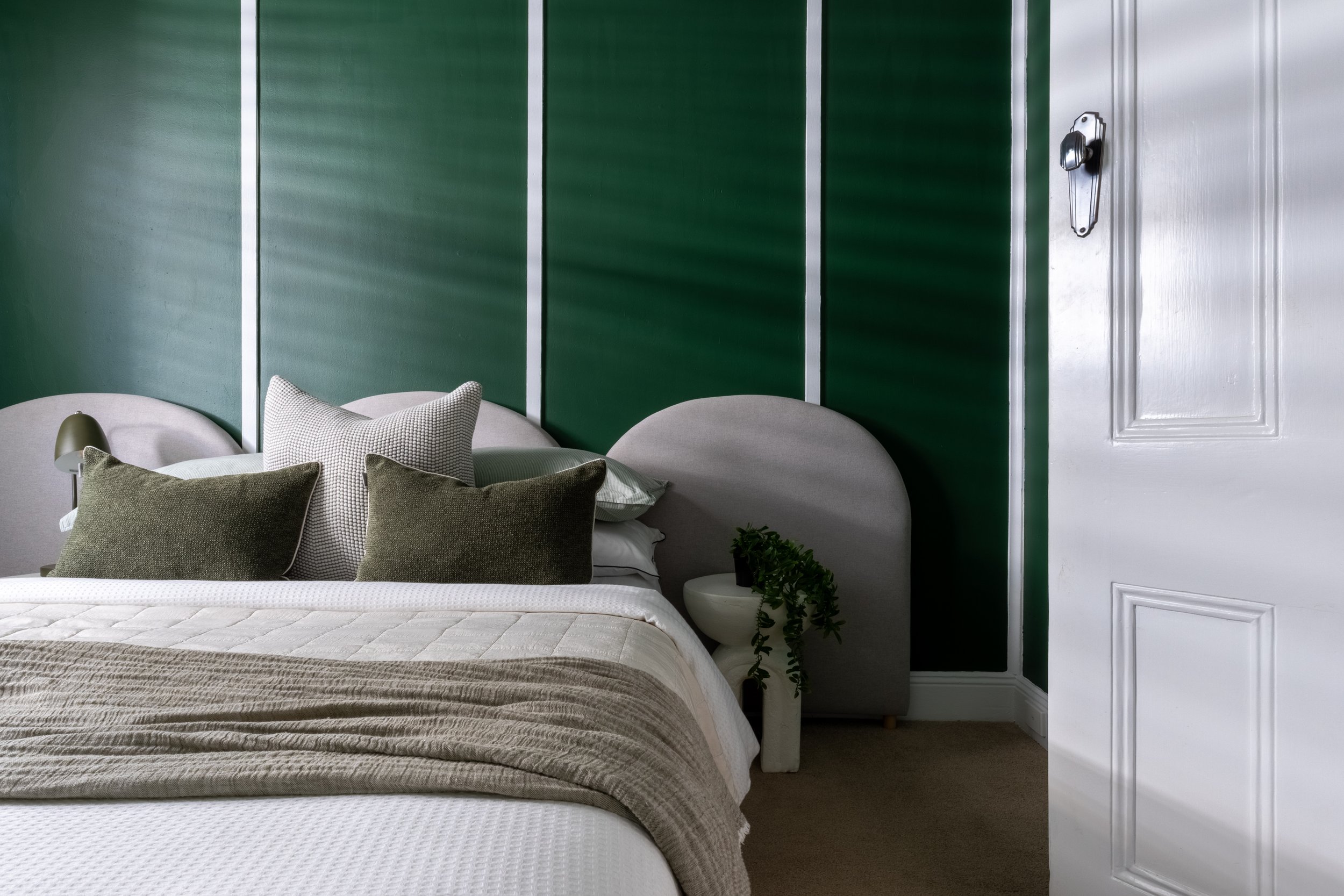 A cozy bedroom with a bed covered in a textured beige blanket, multiple pillows in shades of brown, beige, and white. To the left, a small side table with a dome-shaped green lamp. Behind the bed, there is a green paneled wall with white trim and a w