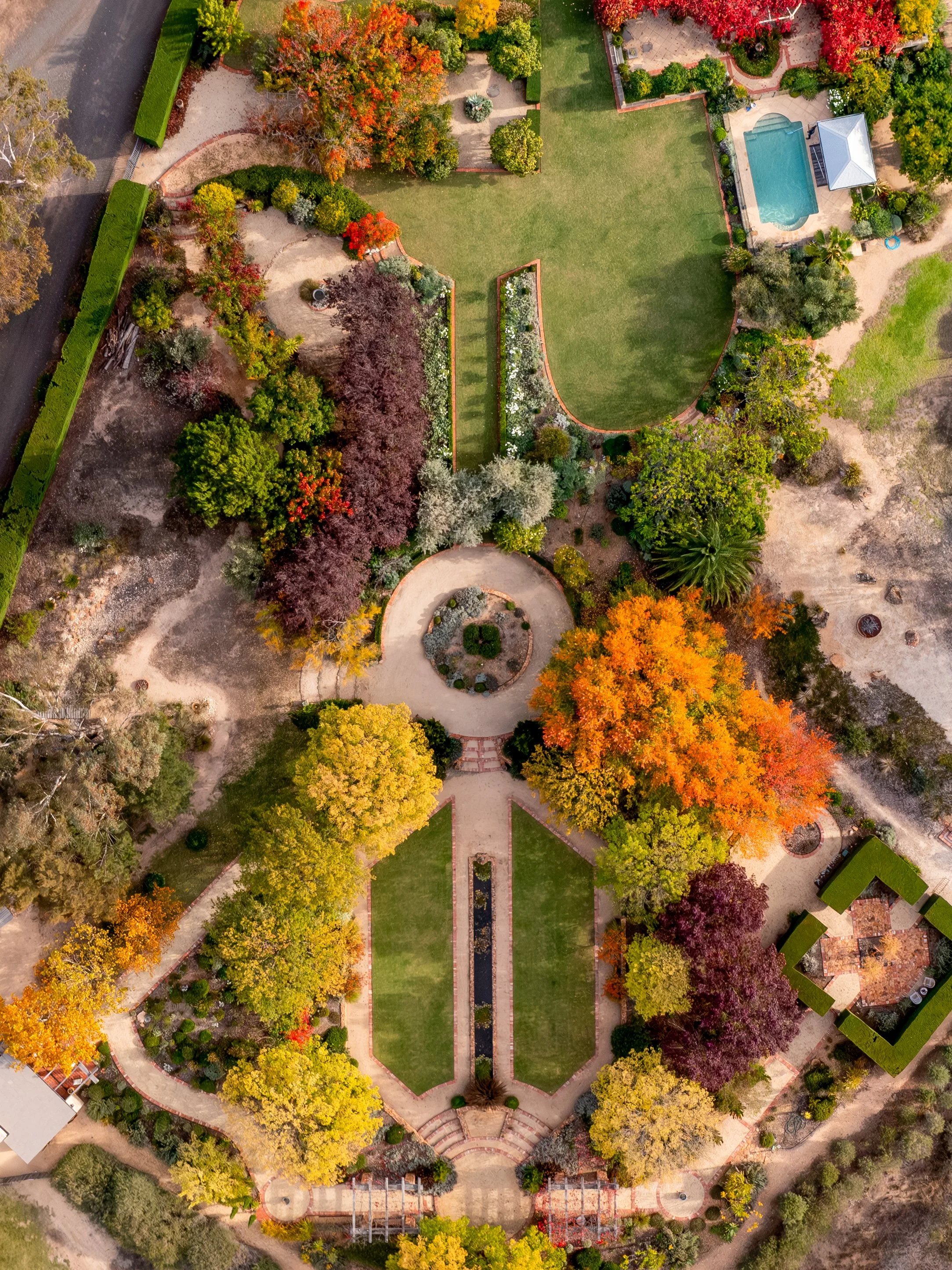 An aerial view of a landscaped park with colorful trees, green lawns, pathways, and a swimming pool with a gazebo nearby.