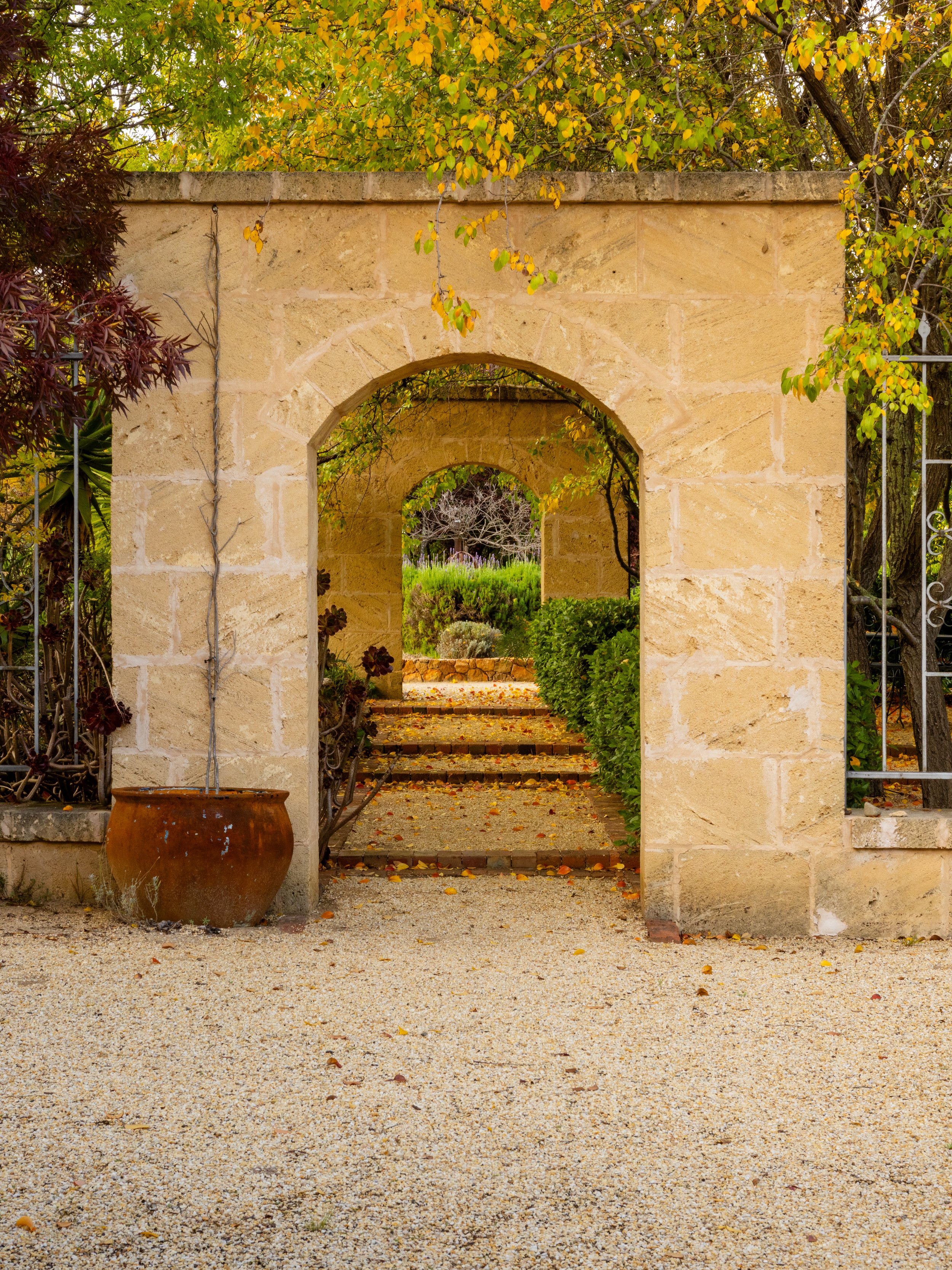 A stone archway leading through a garden with green and yellow foliage, a gravel path, and a large terracotta pot to the left.
