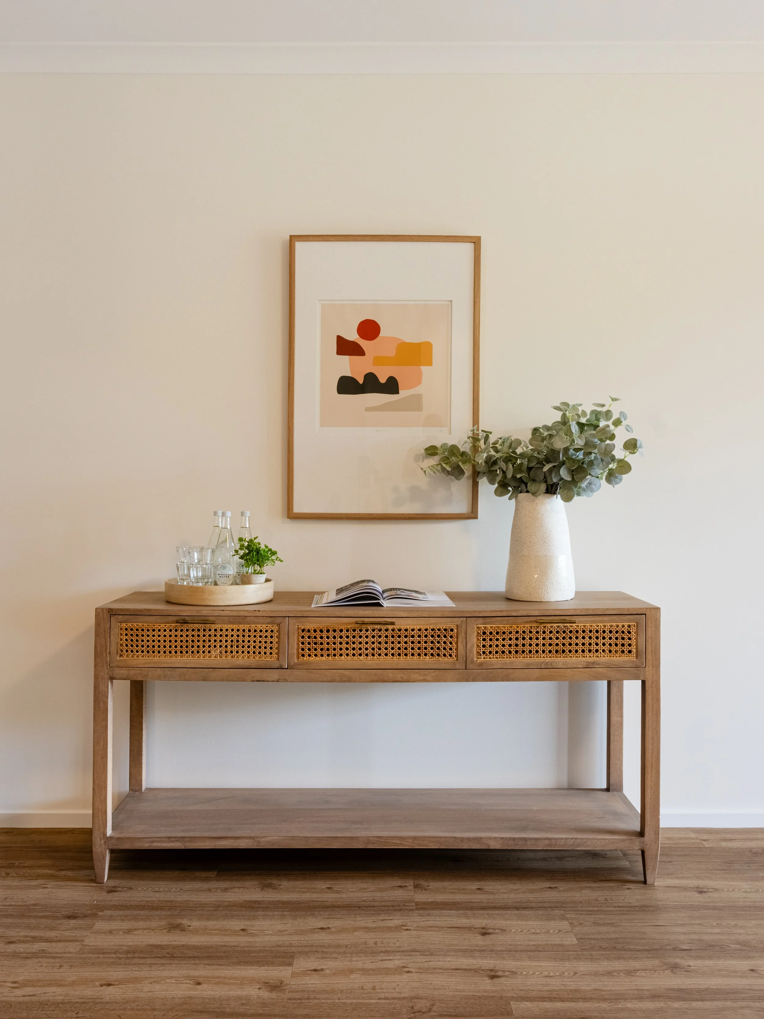 A wooden sideboard with rattan panels, decorated with a tray holding glasses and a small plant, an open book, a tall white vase with green foliage, a framed abstract artwork on the wall, and a wooden floor.