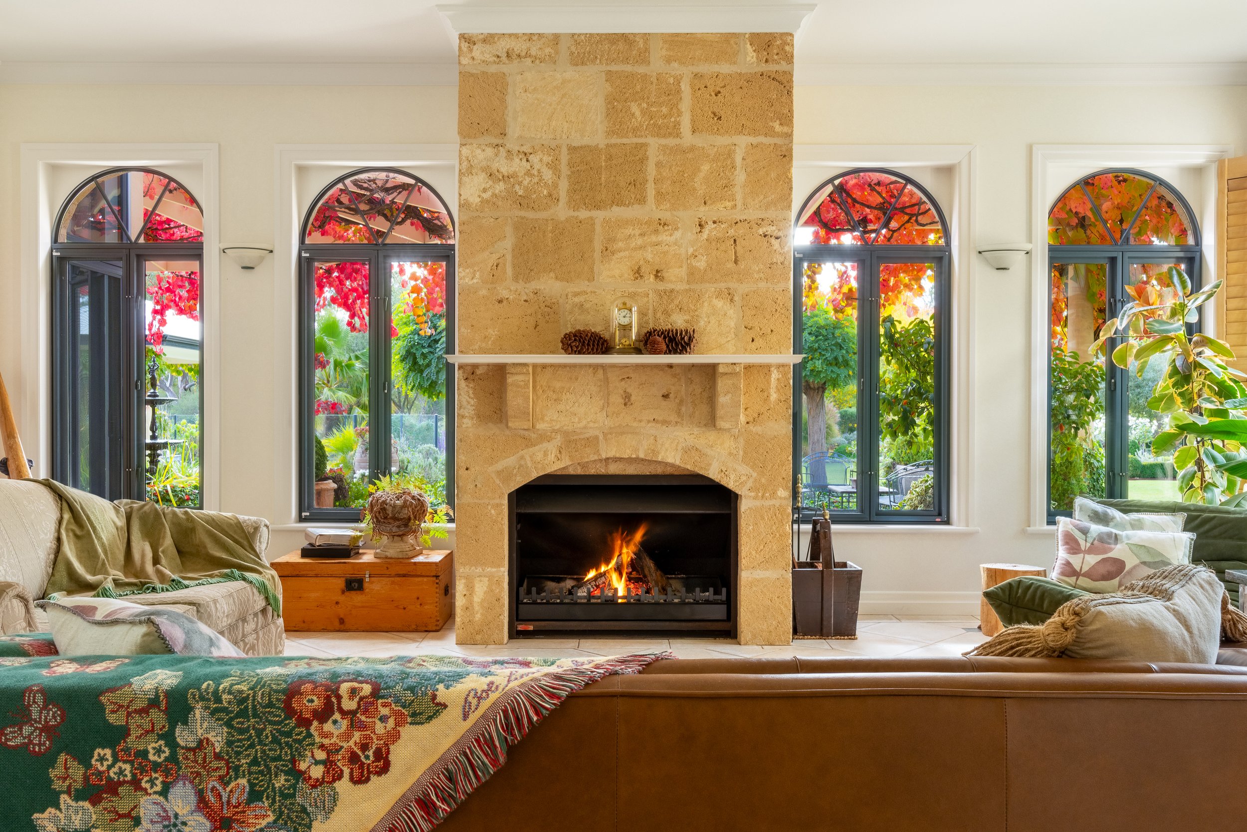 Living room with a stone fireplace, decorated for fall with a fire burning. Four tall arched windows with black frames and colorful fall foliage visible outside. Furniture includes a beige sofa with green and plaid pillows, a wooden chest with decora