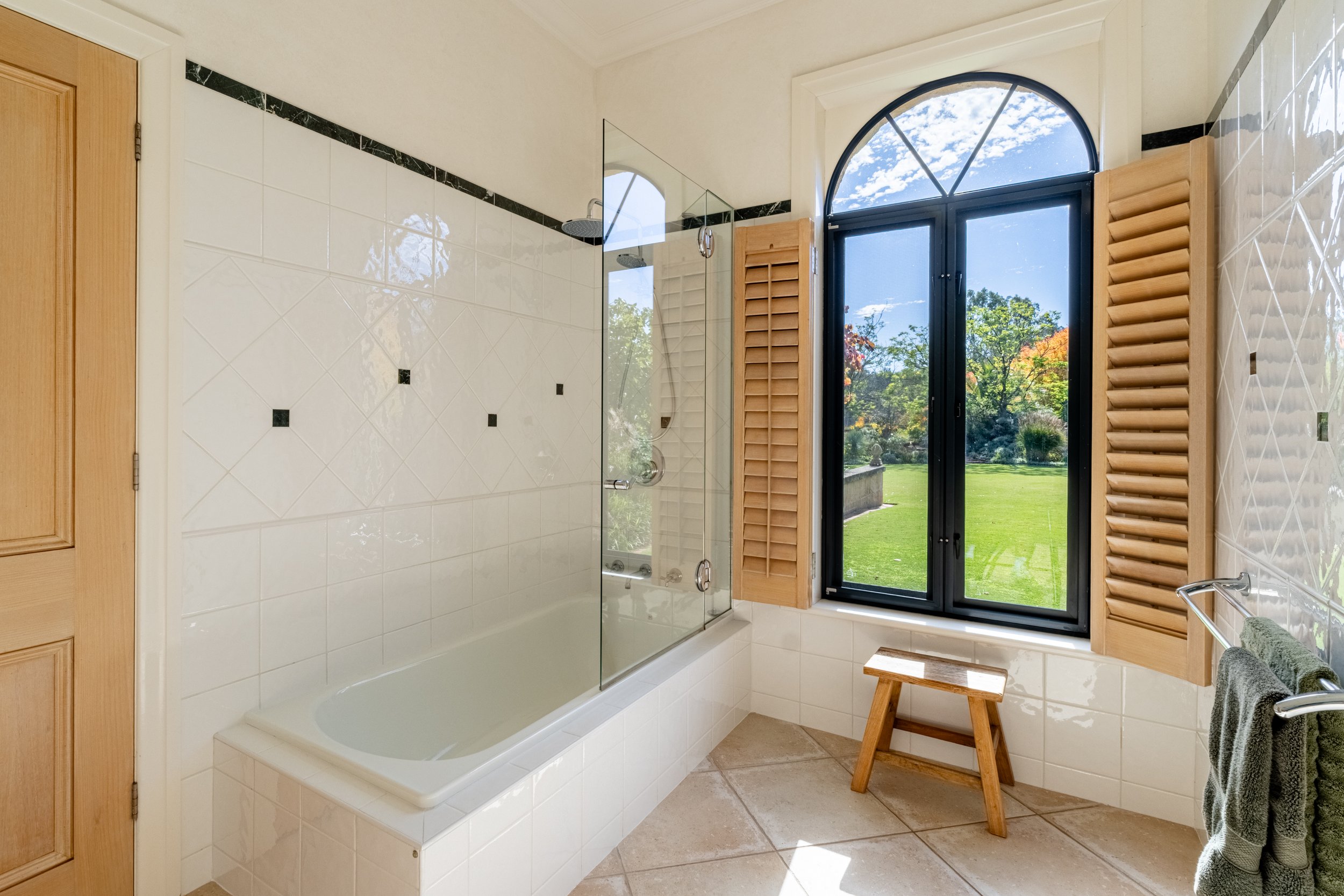 Bathroom with a large window showing a garden outside, a bathtub, a glass shower enclosure, a wooden stool, and towels.