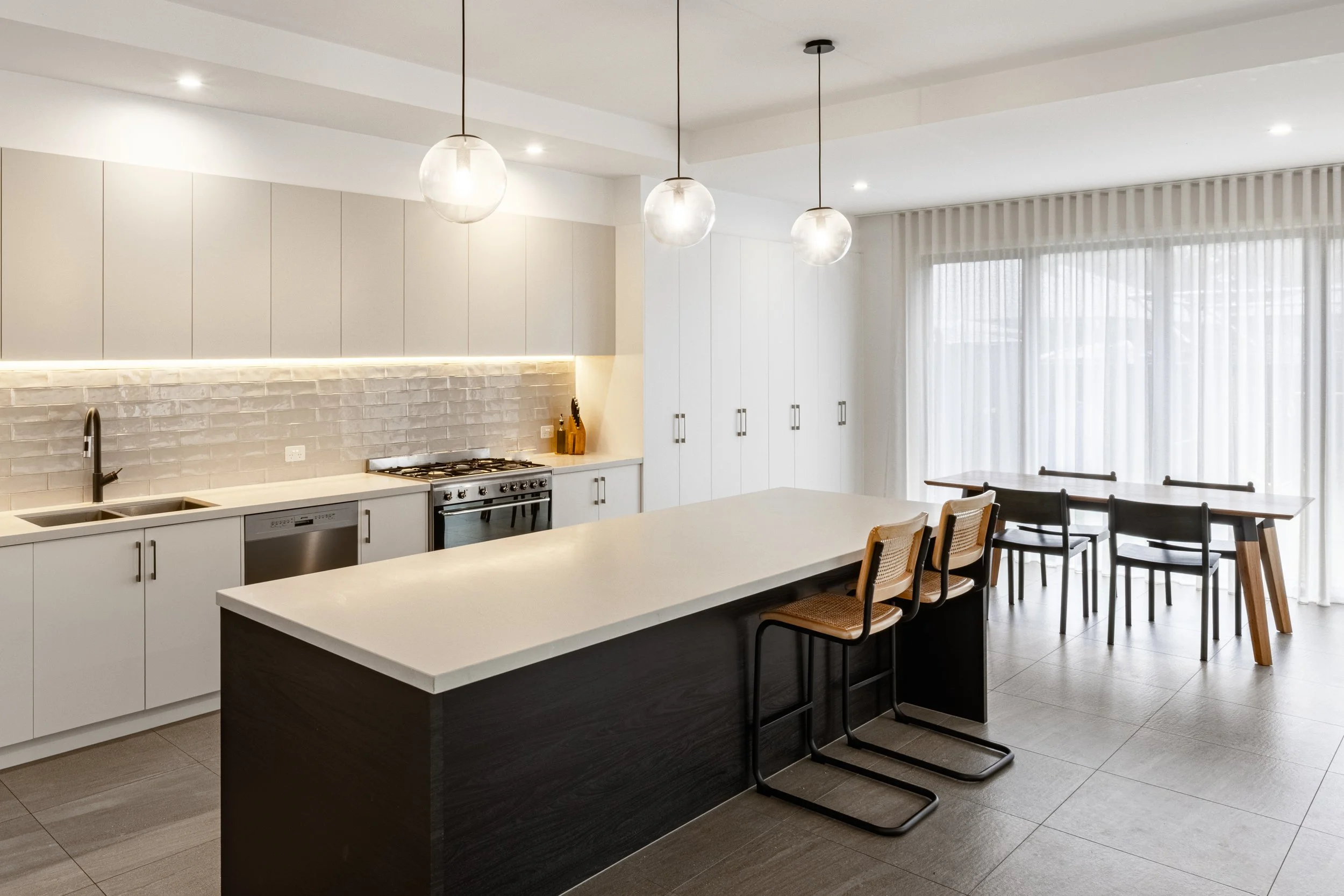 Modern kitchen with white cabinets, a white kitchen island, pendant lights, a gas stove, and a dining area with a table and chairs near large windows with sheer curtains.