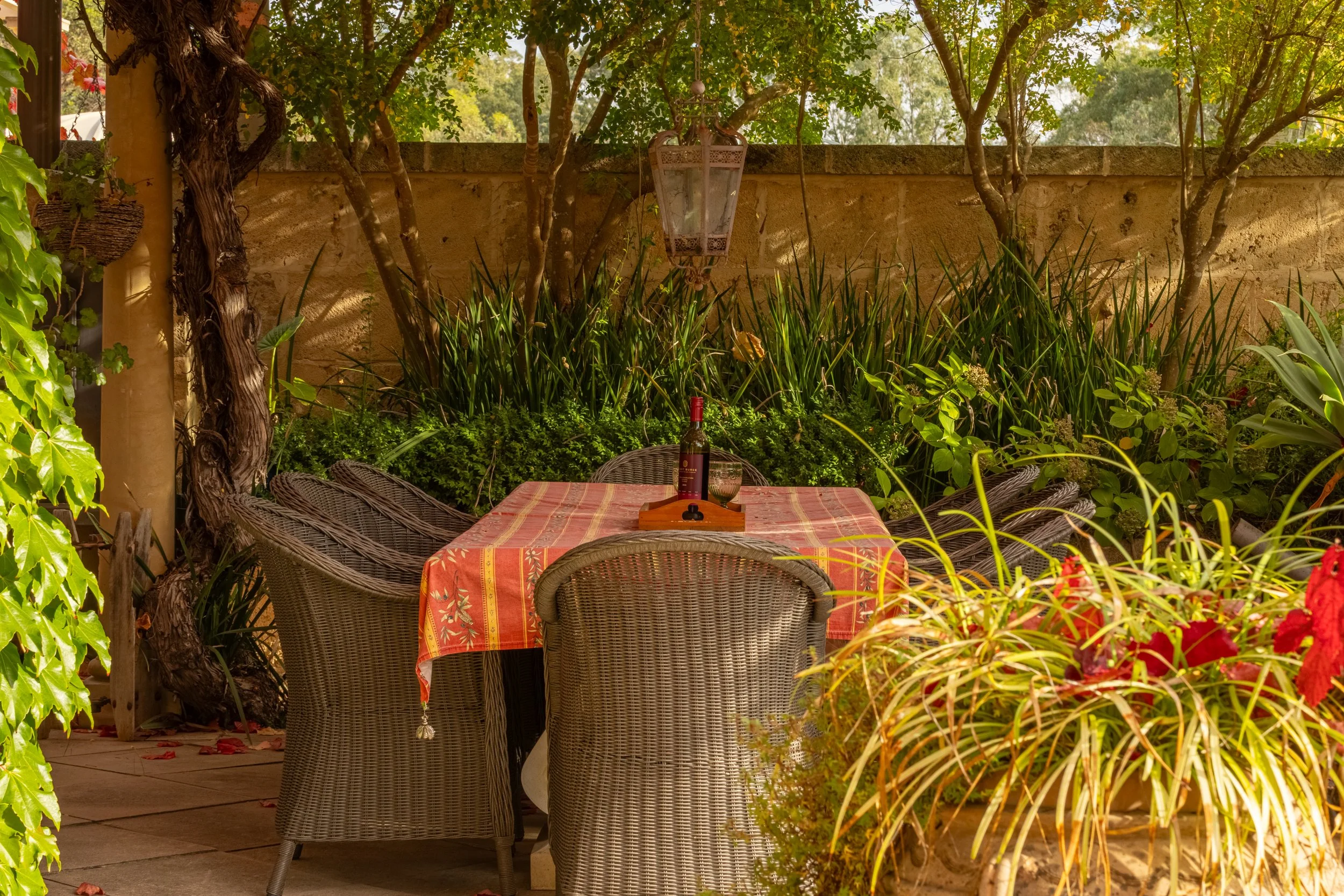 A cozy outdoor dining area with a table covered by an orange and yellow patterned tablecloth, surrounded by wicker chairs. There is a bottle of wine, a wine glass, and a small wooden serving tray on the table. The setting is lush with greenery, trees
