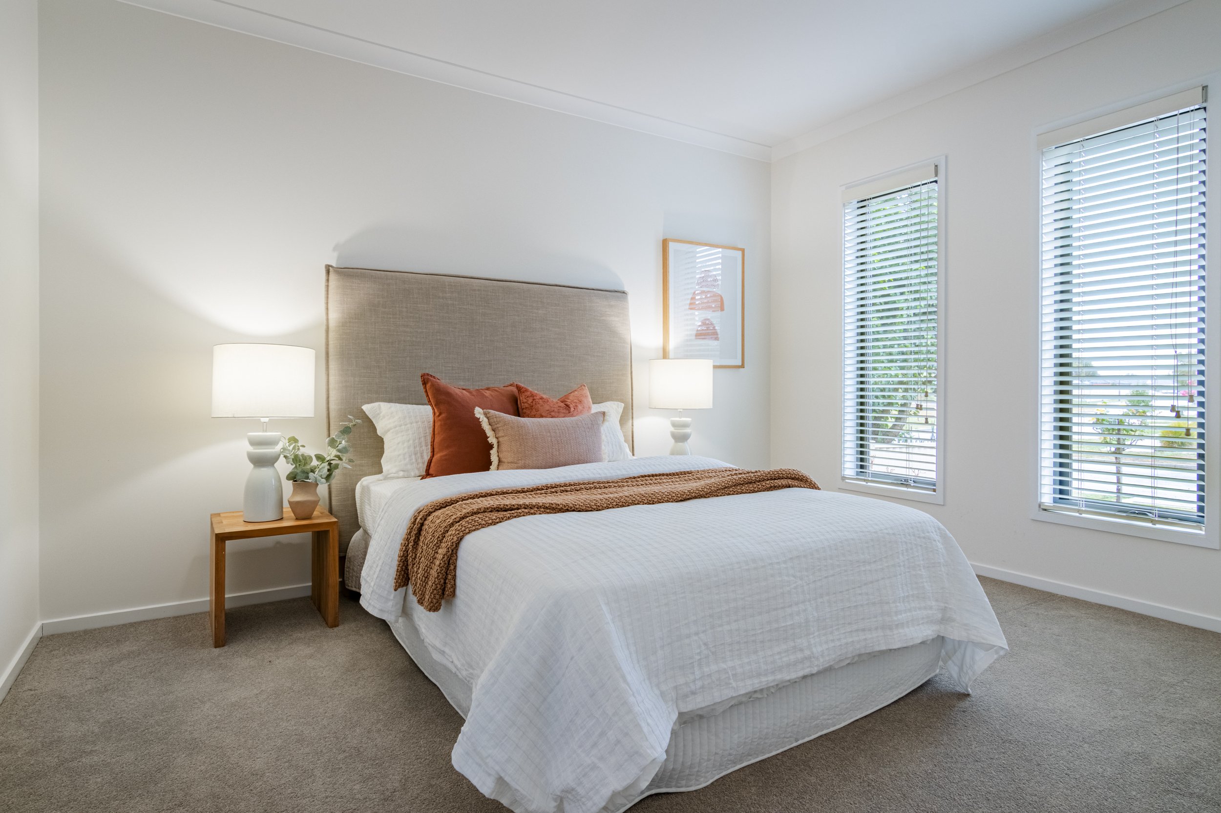 A neatly made bed with white bedding, orange and beige pillows, a beige headboard, beside two white lamps on wooden nightstands, framed artwork on the wall, and two windows with white blinds, in a bright bedroom.