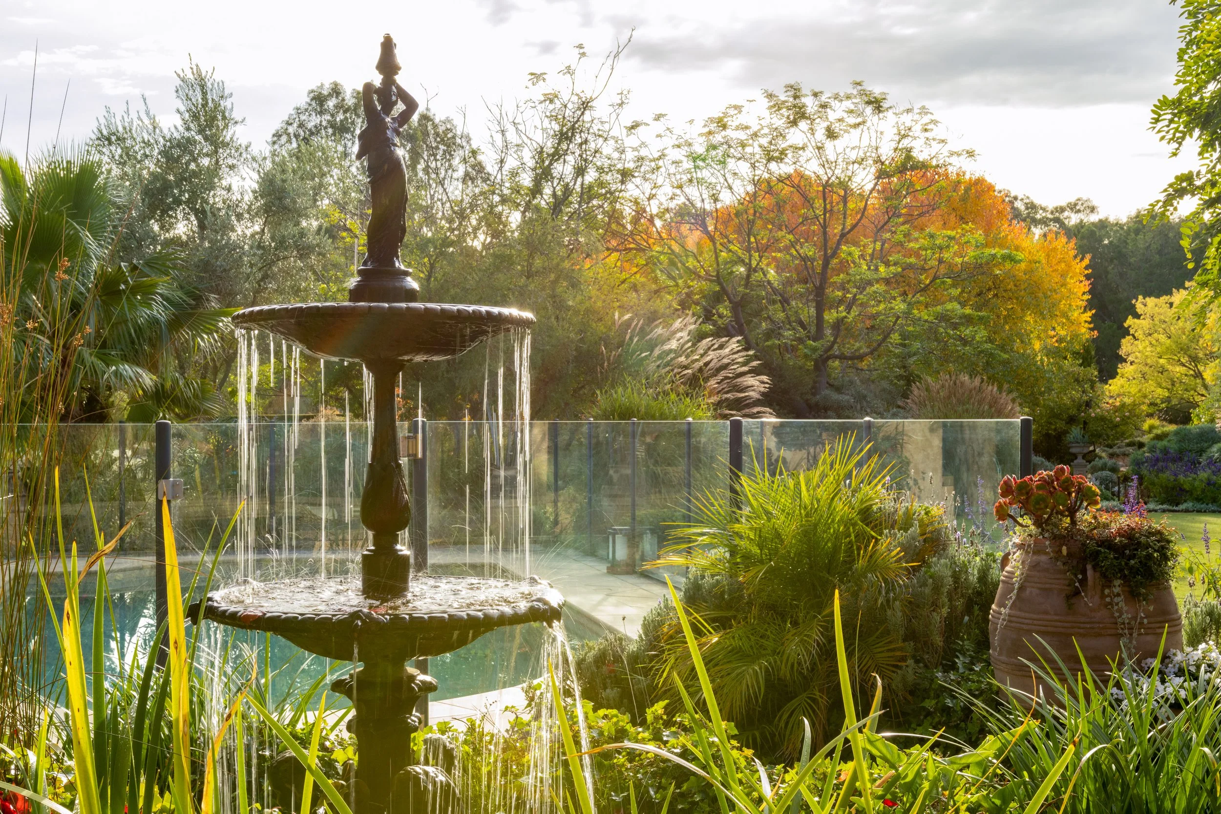 A garden scene with a multi-tiered fountain surrounded by various plants and trees. Autumn trees with orange and yellow leaves are in the background, and the sky is partly cloudy with sunlight filtering through.