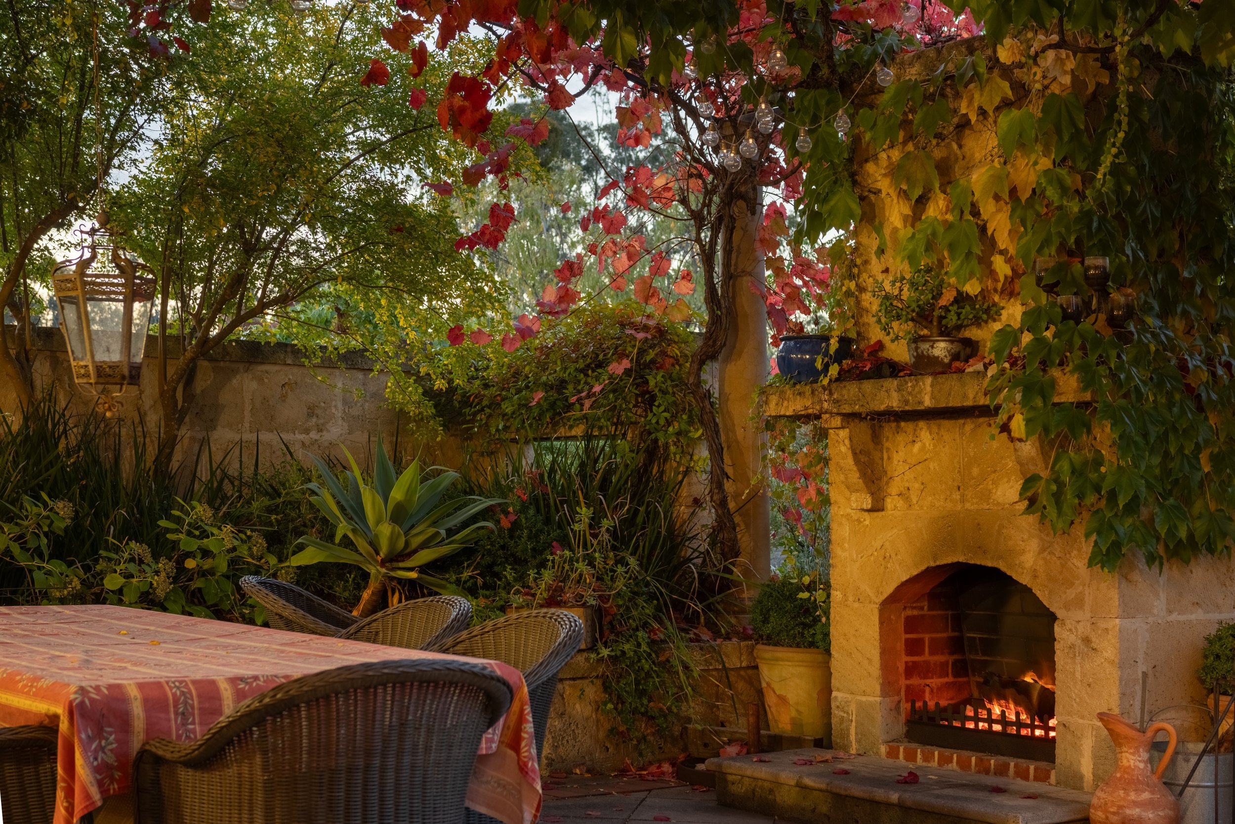 A cozy outdoor patio with a stone fireplace, surrounded by green and red climbing plants, lit by string lights, with wicker chairs and a table with an orange patterned tablecloth.