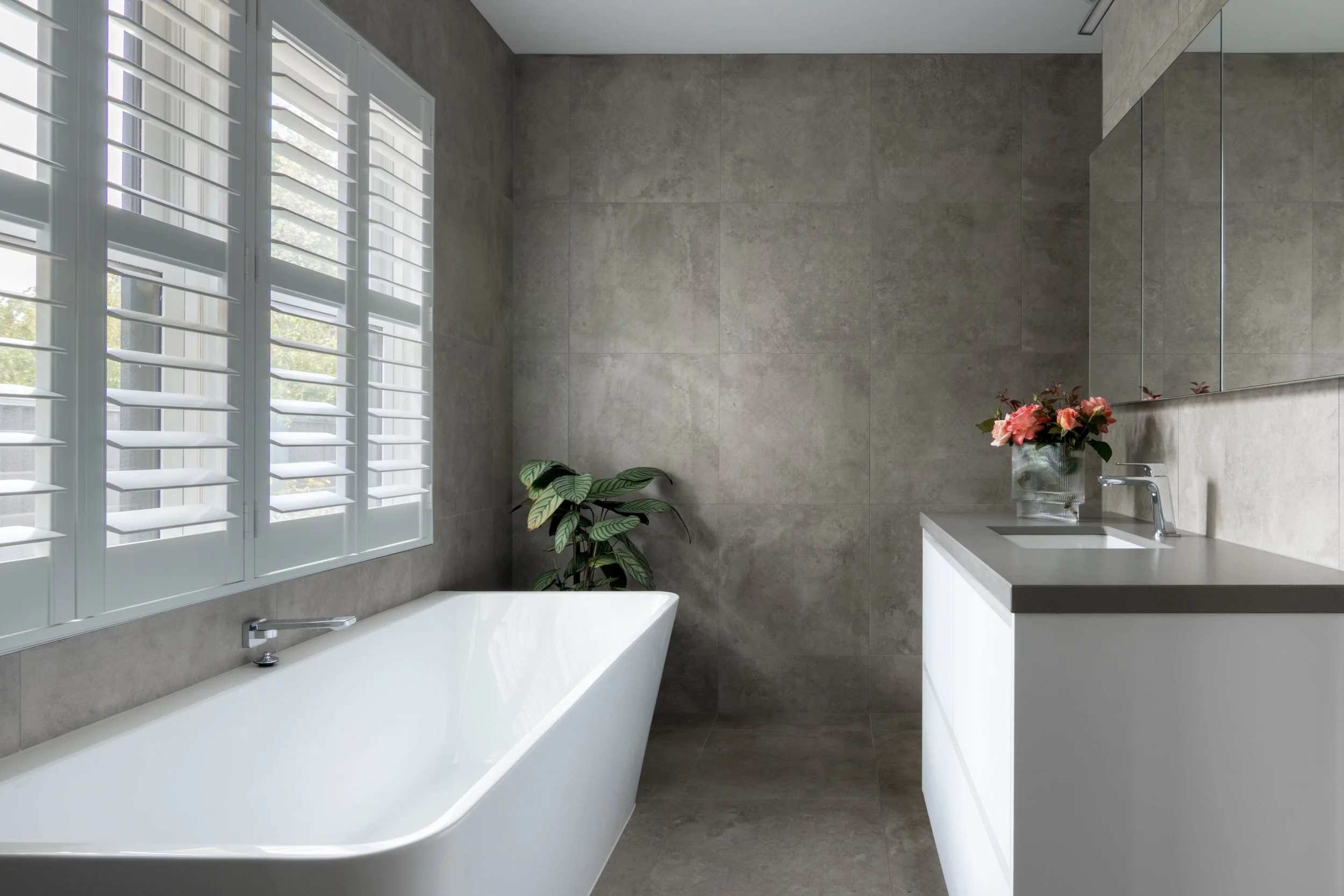 Bathroom with large window with white shutters, gray tiled walls, a white bathtub, a small potted plant with large green leaves, a vanity with a sink, and a vase with pink flowers.