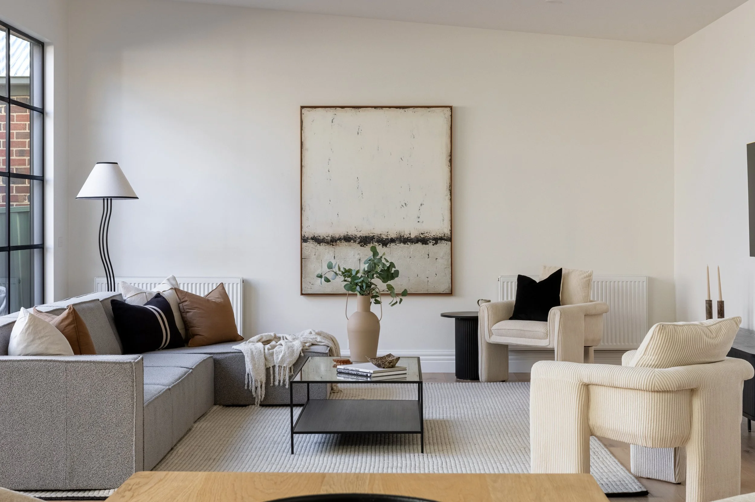 Living room with a beige sofa, black coffee table, cream armchair, wall art, window, and a floor lamp.