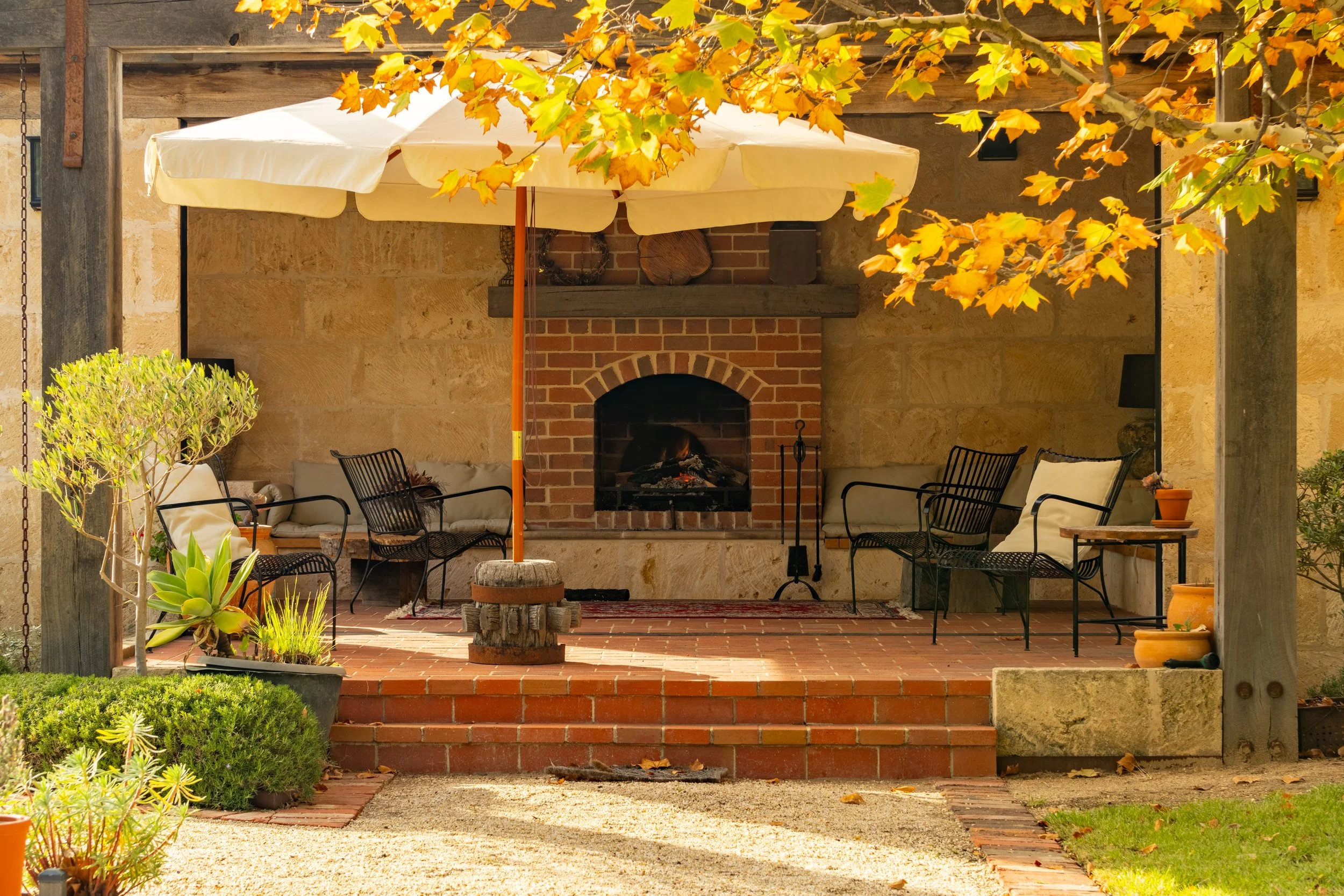 Outdoor patio area with a brick fireplace, surrounded by chairs with cushions, a white umbrella, and autumn leaves overhead. Plants and greenery decorate the space, with steps leading up to the patio.