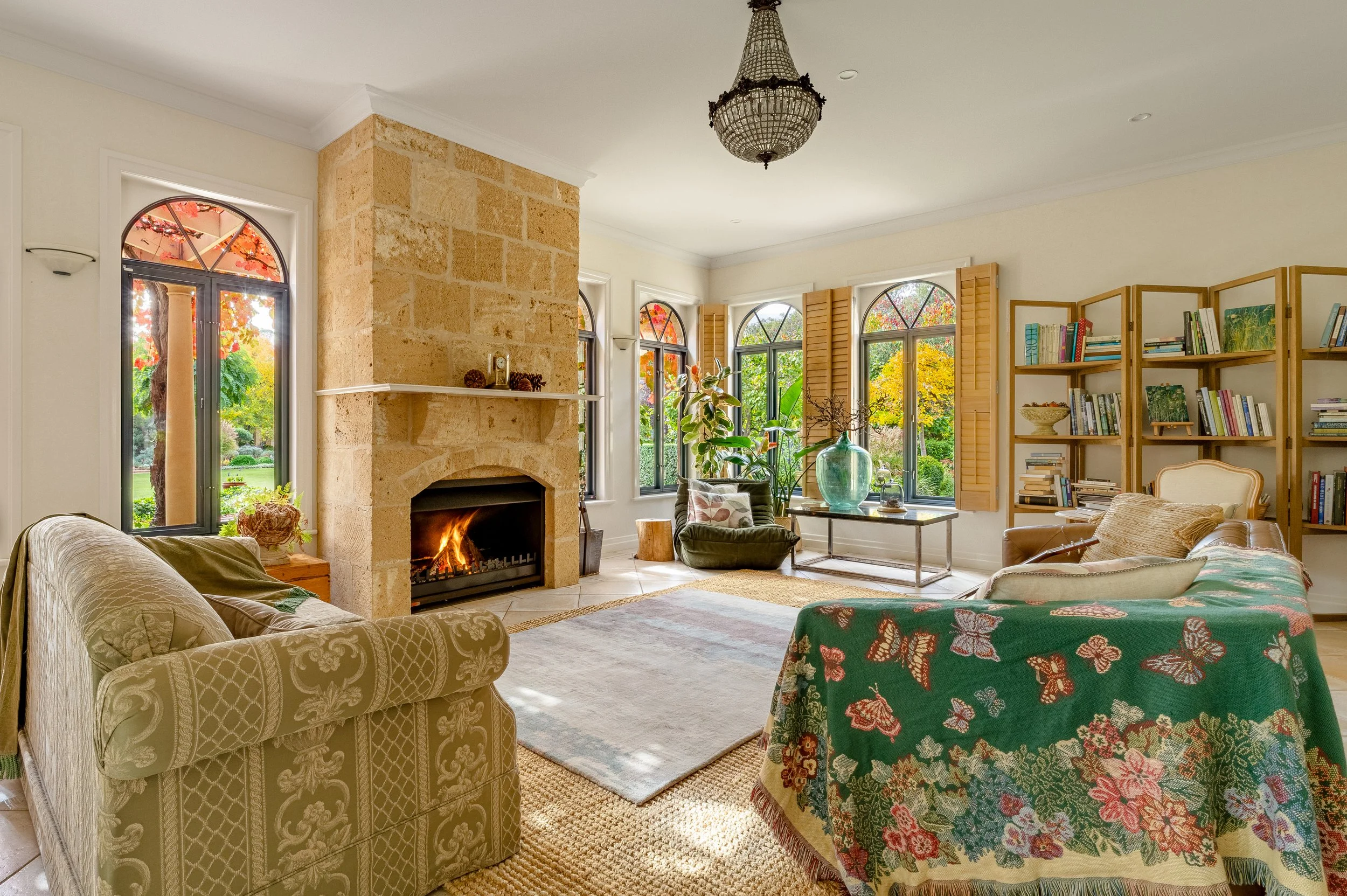 Living room with stone fireplace, surrounded by beige walls, and large windows with wooden shutters showing trees with fall foliage. There are various chairs and a sofa with decorative pillows, a large green vase, a bookshelf, and a chandelier hangin
