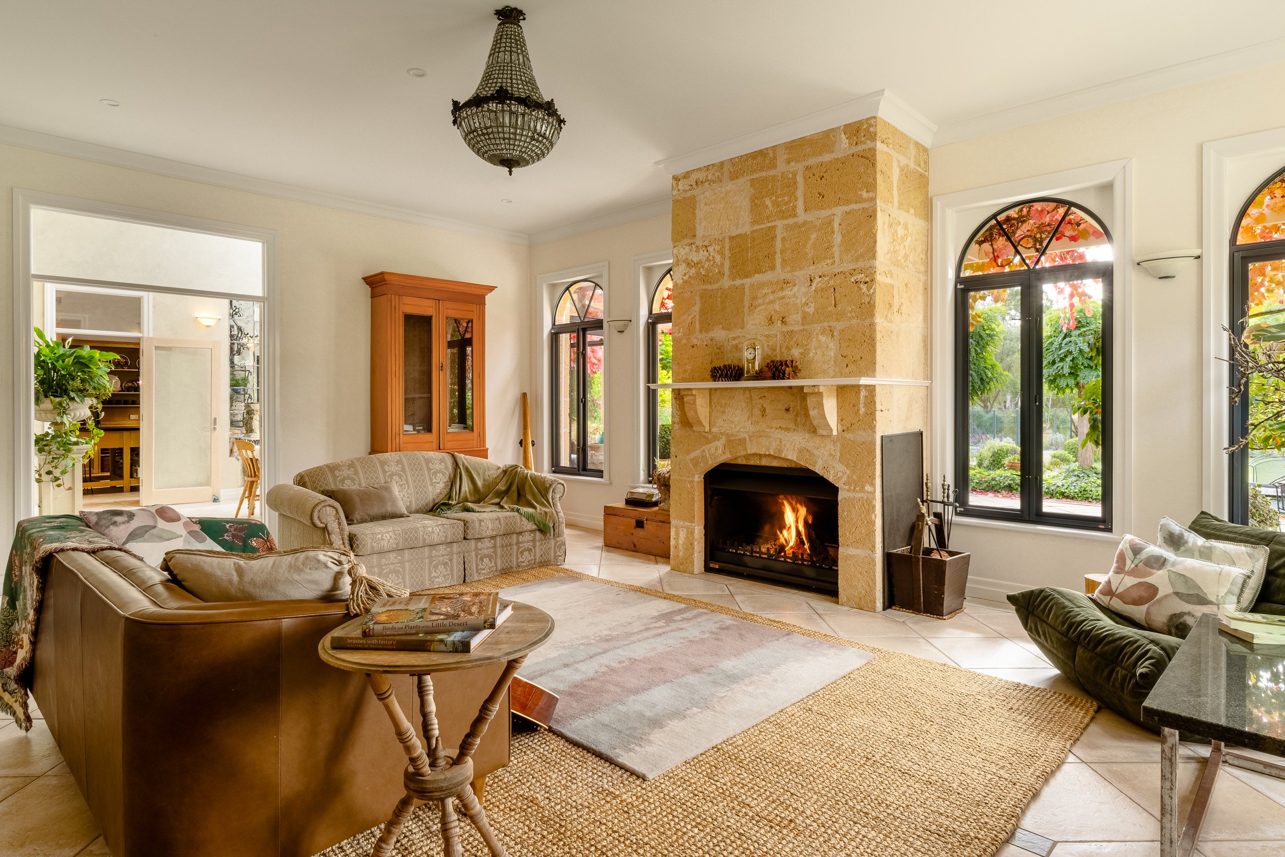 Living room with a stone fireplace, sofa, armchairs, large windows with views of trees, and various decorative items.
