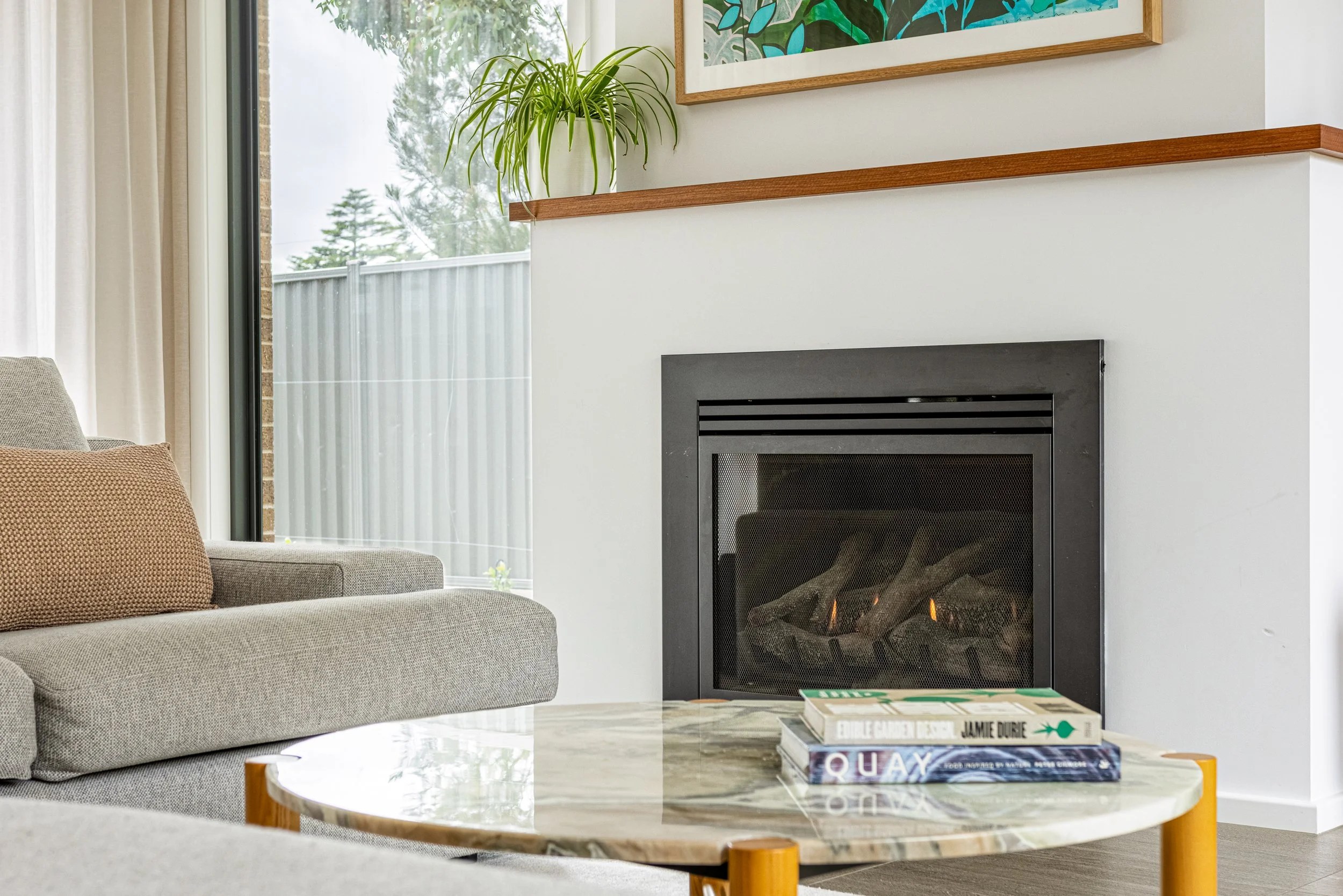 Modern living room with a gray sofa, a marble-topped coffee table holding books, a fireplace, and a large window with white curtains, overlooking trees outside.