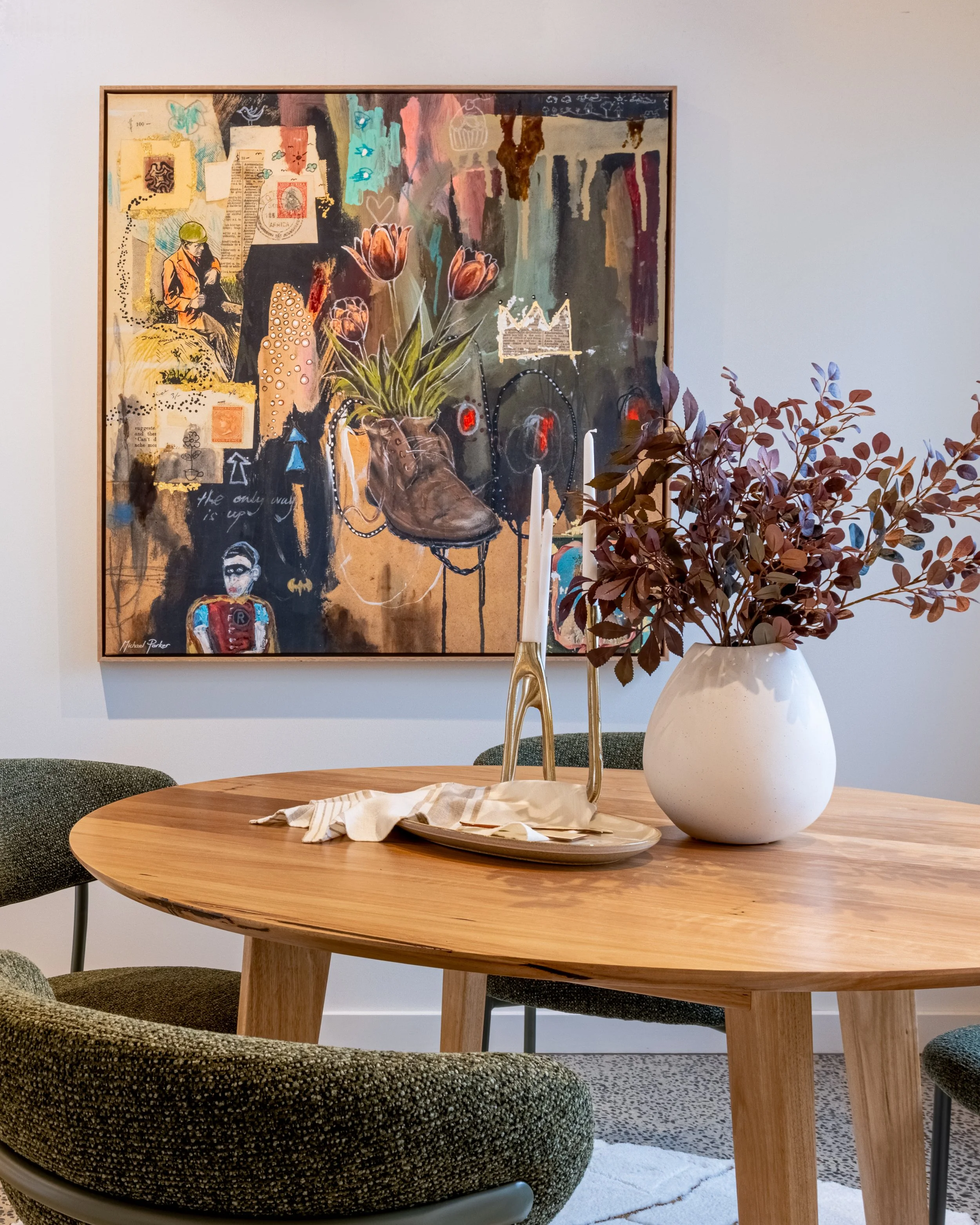 A decorated round wooden dining table with a white vase containing dark purple leaves, a tall gold candlestick, and a beige plate with a cloth napkin. In the background, a large abstract collage painting on the wall and gray upholstered chairs surrou