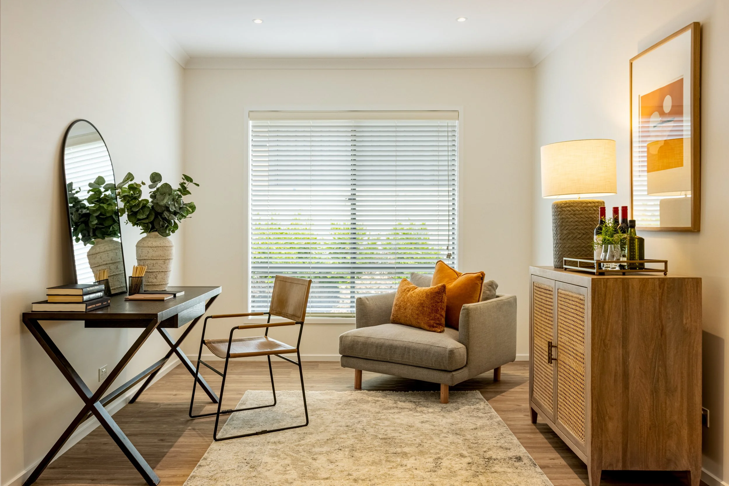 A cozy living room with a large window with white blinds, a beige armchair with an orange pillow, a wooden cabinet with decorative bottles, a table with a green plant, a mirror, and a lamp, all in warm neutral tones.