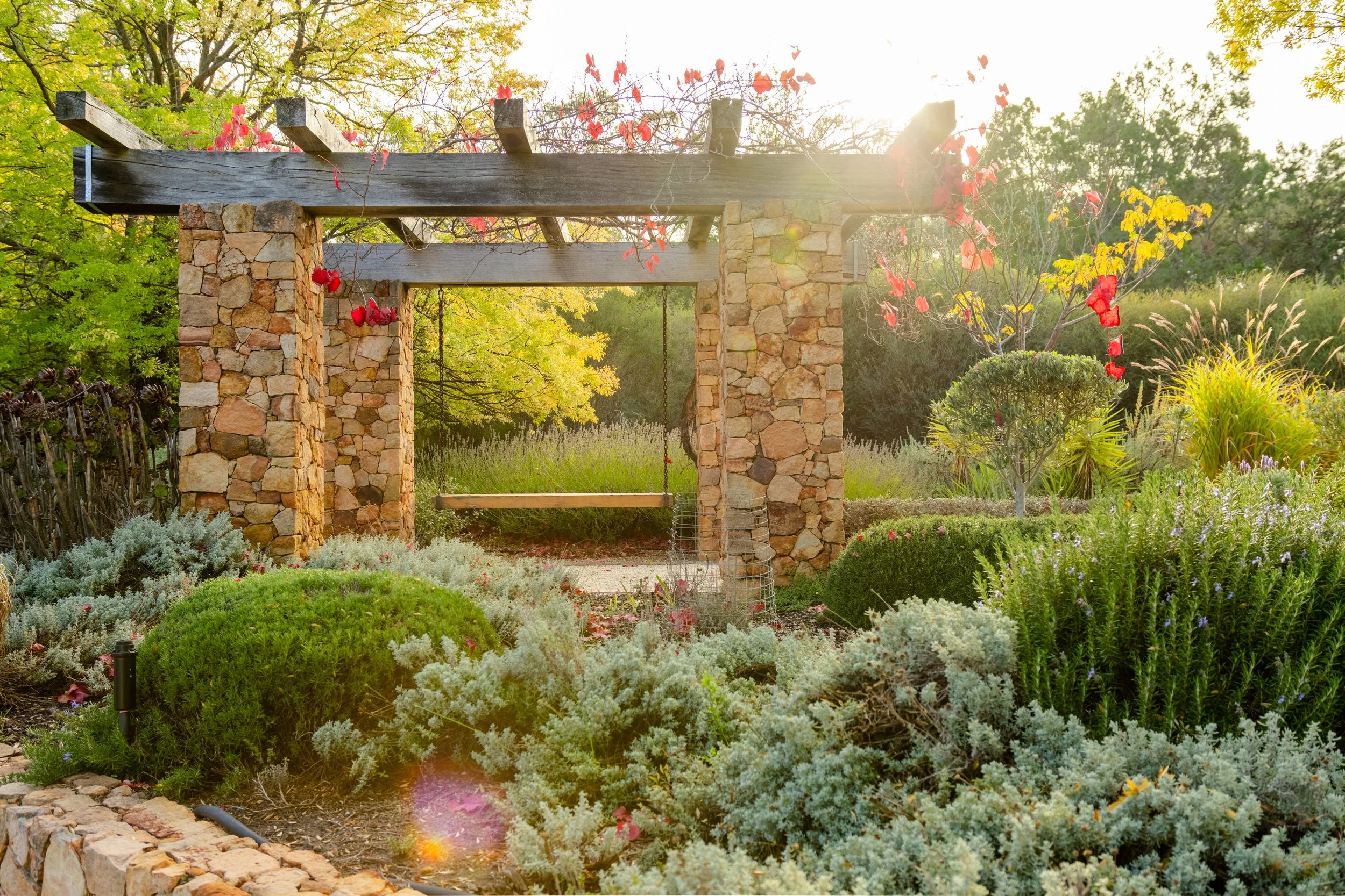 A garden scene featuring a stone and wood pergola surrounded by lush green foliage, flowering plants, and shrubs, with sunlight filtering through the trees.