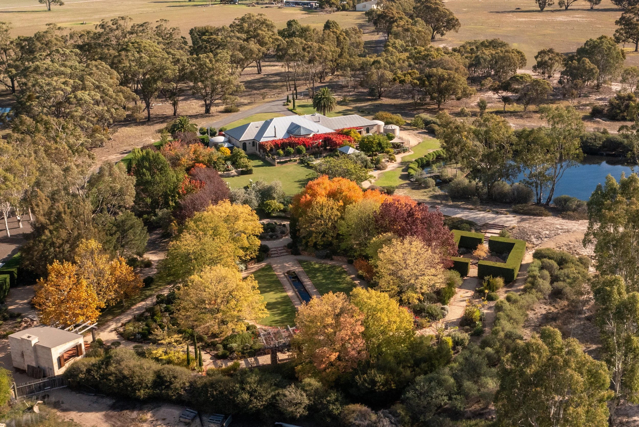 An aerial view of a large house with a gray roof, surrounded by colorful autumn trees, a manicured lawn, a pond, and a road in a rural area with open fields and scattered trees.