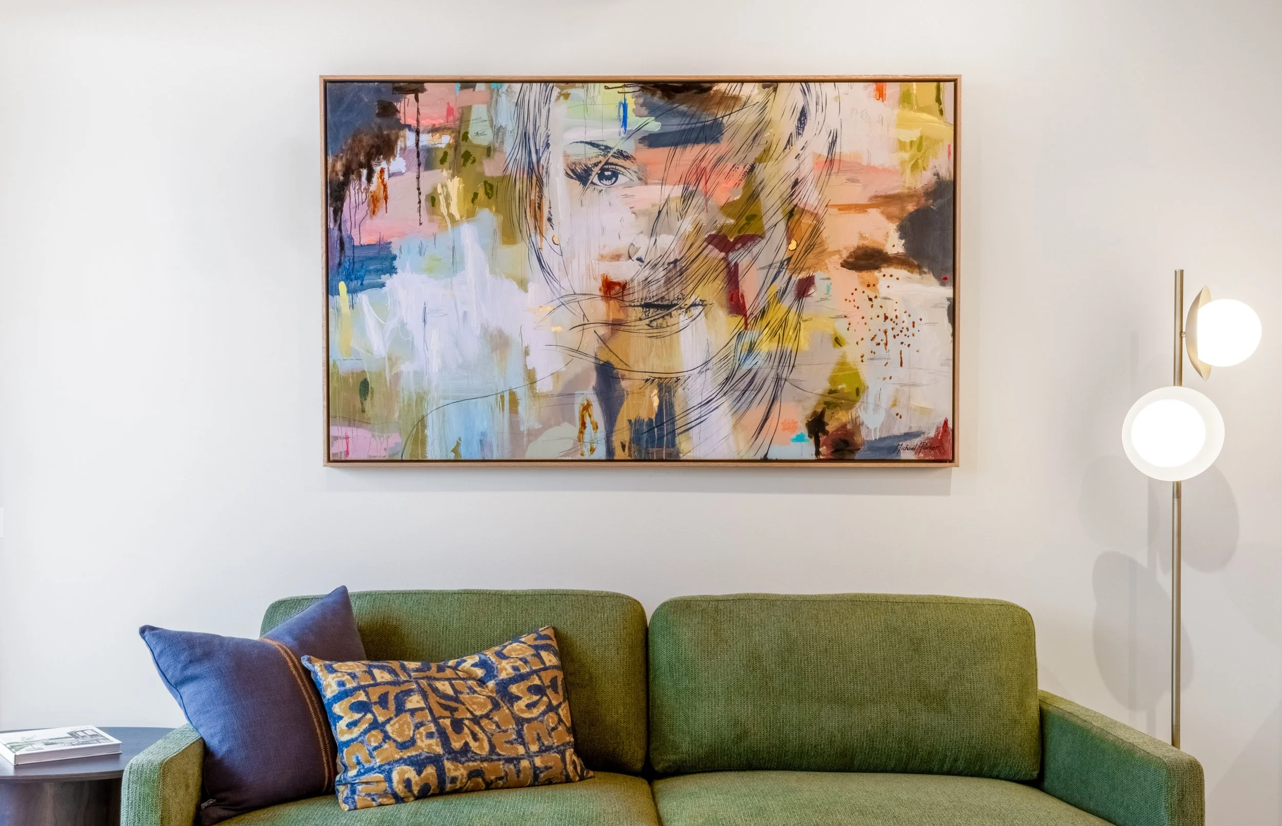 Large modern abstract painting on white wall behind a green sofa with blue and patterned pillows, adjacent to a standing lamp with two glowing circular lights.