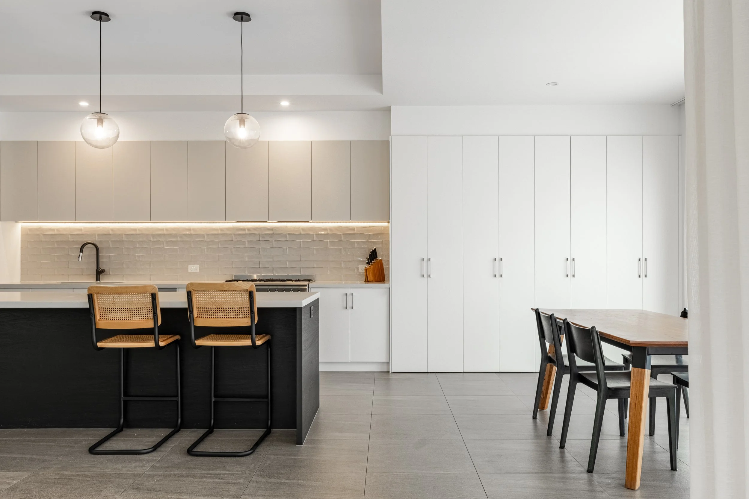 Modern kitchen and dining area with minimalist design, white cabinets, a kitchen island with woven rattan chairs, pendant lighting, and a brick backsplash.